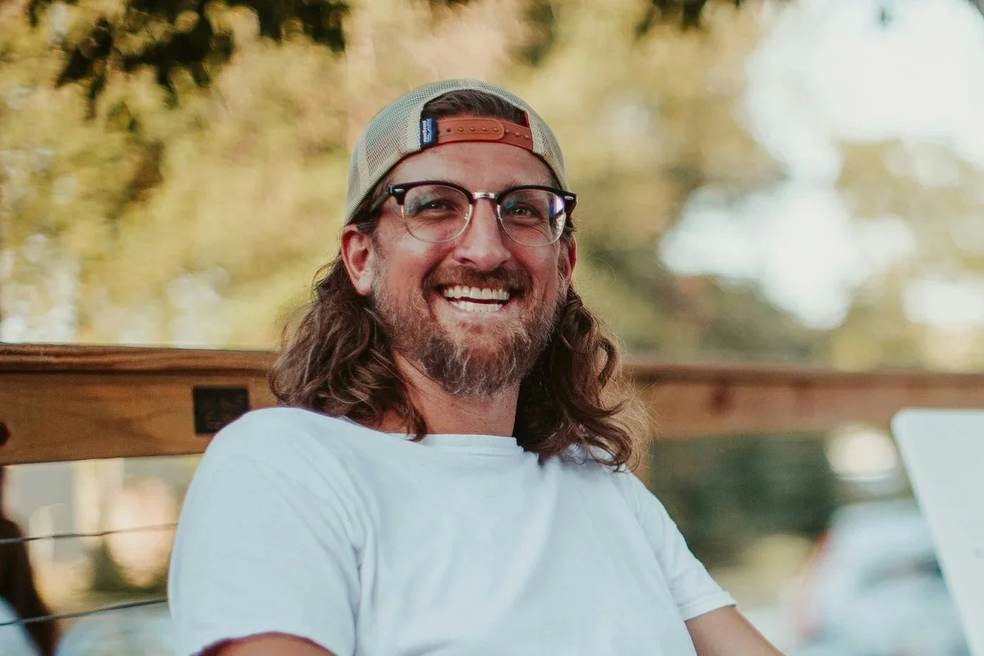 A man with long hair, beard, eye glasses, wearing a baseball cap and a white t-shirt, smiling while sitting outdoors