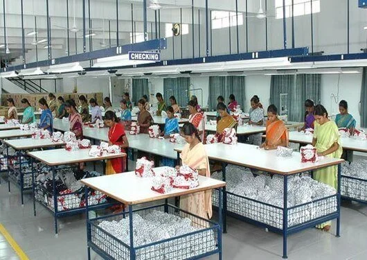 Women working with embroidered fabrics at tables in a large factory or workshop.