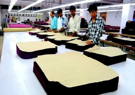 Five individuals working in a textile factory, cutting large fabric sheets on a white table with industrial lighting overhead.