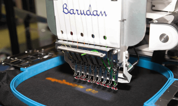 Close-up of a computer embroidery machine stitching the words 'Goodbye' on black fabric with orange and gray threads, with a blue embroidery hoop holding the fabric in place.