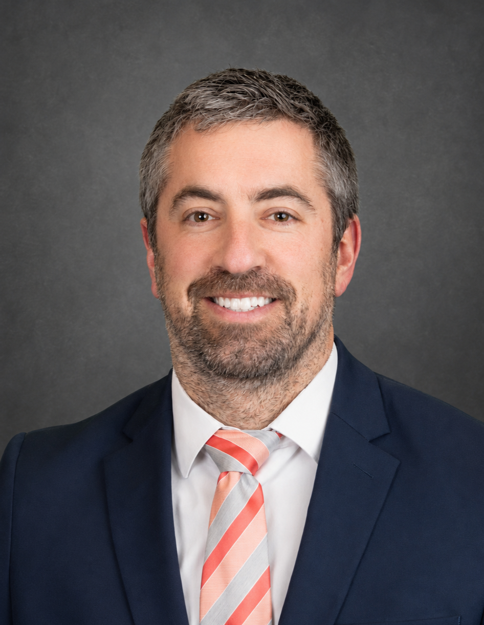 Professional headshot of a middle-aged man with short gray hair and a beard, wearing a navy suit, white shirt, and a pink and white striped tie, smiling against a gray background.