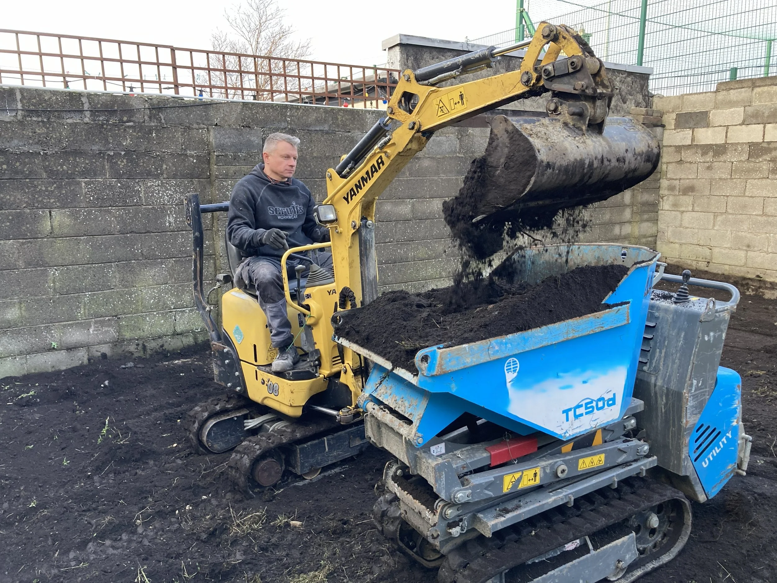 A man operating a small yellow excavator tractor, filling a blue and gray soil screener with dark soil at a construction site, with concrete walls and fencing in the background.
