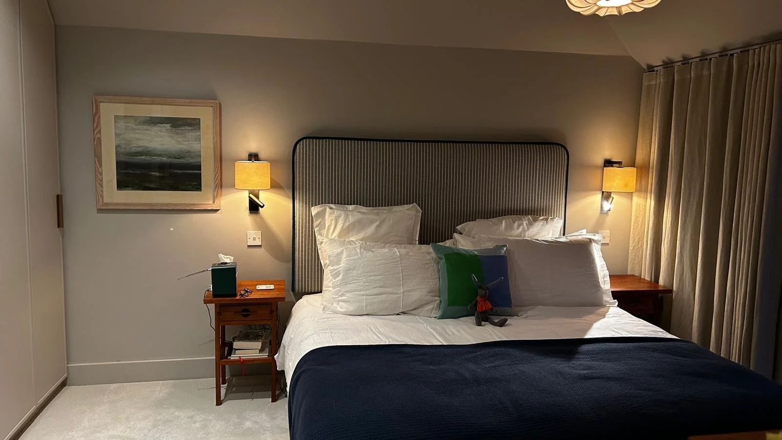 A neatly made hotel bedroom with a large bed, two pillows, a stuffed toy, and a dark blue blanket. Two wall-mounted lamps flank a striped headboard, with a wooden nightstand on each side, one holding a tissue box, glasses, and a remote. There's artwork on the wall and long curtains to the right.