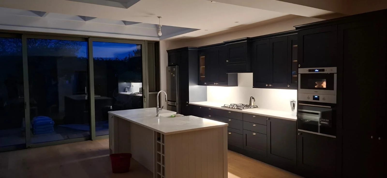 Modern kitchen with black cabinets, white countertop, kitchen appliances, and sliding glass doors leading outside during evening.