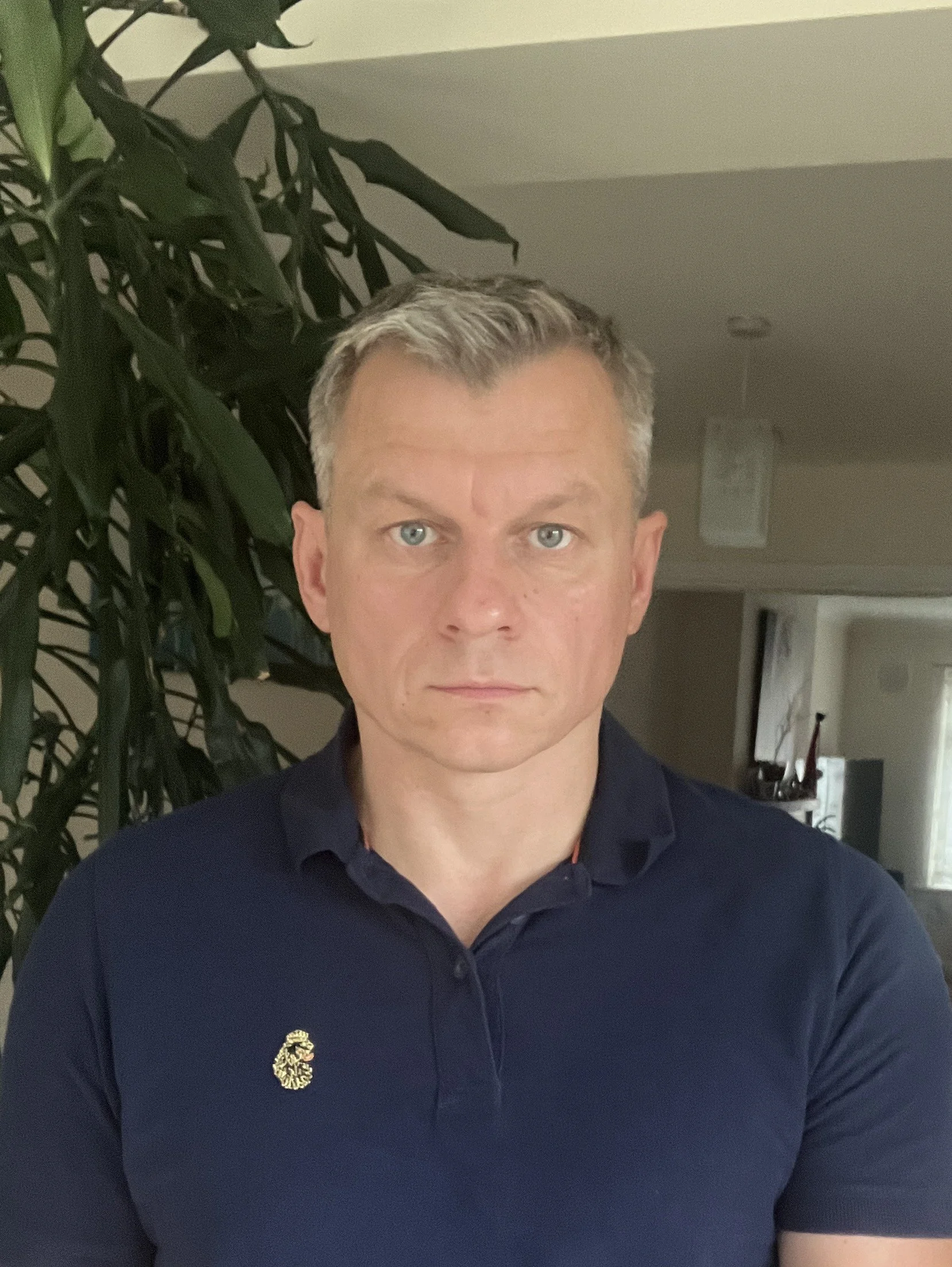A man with short, light-colored hair and blue eyes wearing a navy polo shirt with an embroidered logo of a seahorse on the left chest, standing indoors near a tall plant with broad leaves, with a neutral expression.