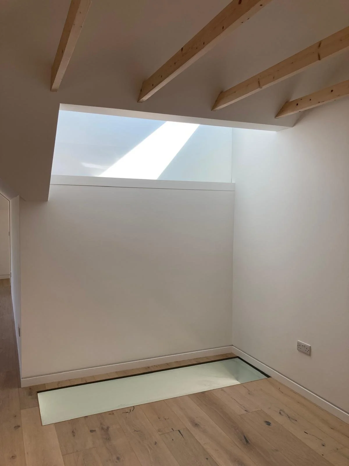 Interior of a room with a skylight, wooden beams on the ceiling, white walls, a wooden floor, and a floor-level glass window with a light-colored border.