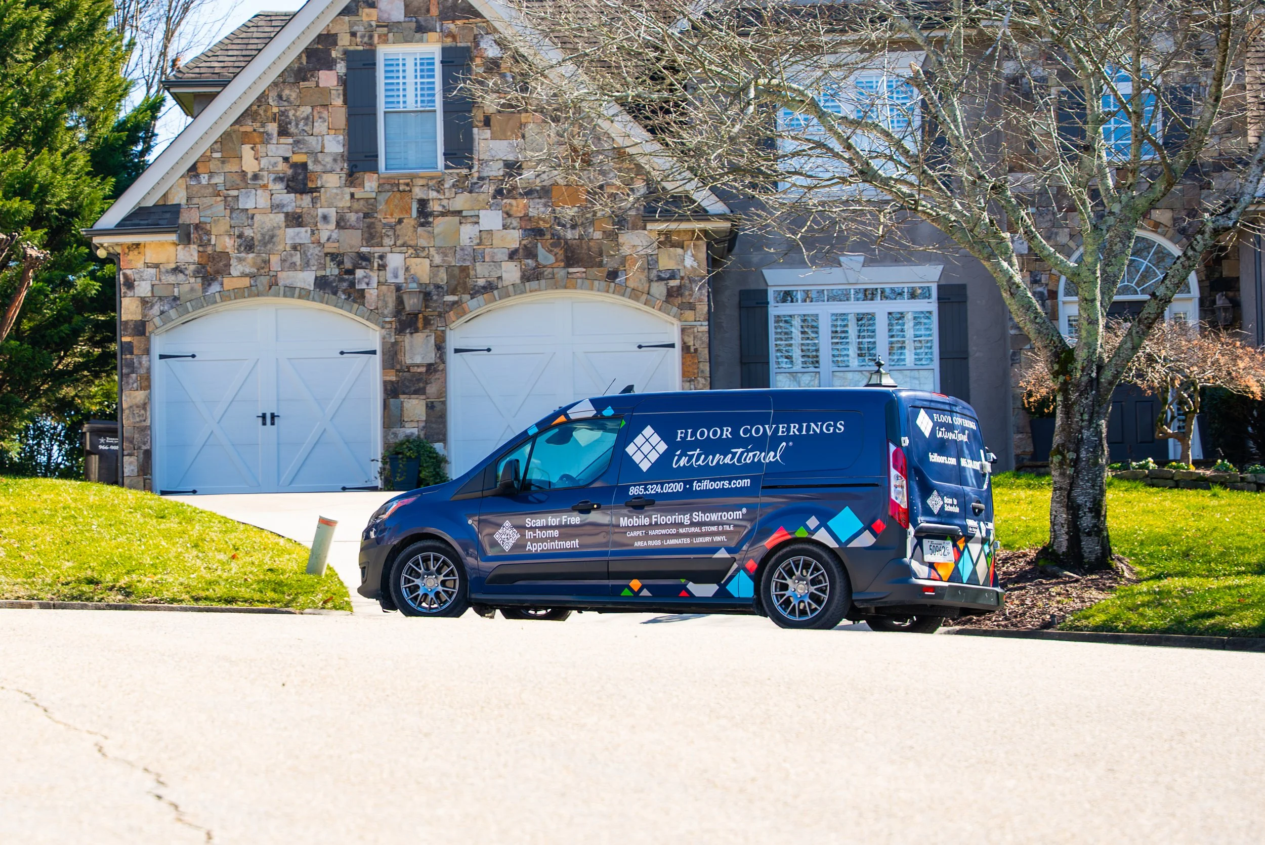 A blue van parked in front of a two-story house with a stone facade, white garage doors, and a large front window with shutters. The van advertises a flooring company and is situated on a driveway with a grassy lawn and a leafless tree nearby.