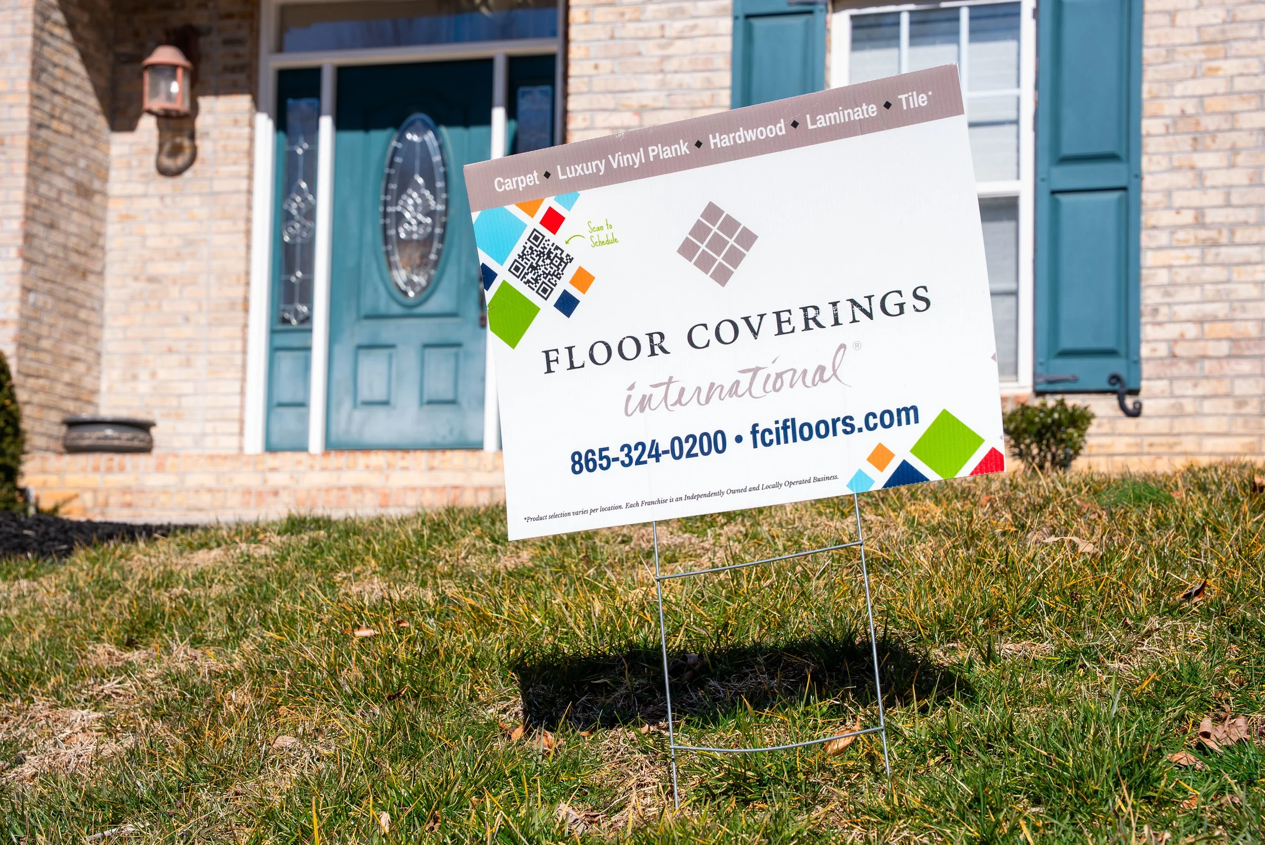 A yard sign advertising flooring options, placed in front of a brick house with a blue front door and window shutters.
