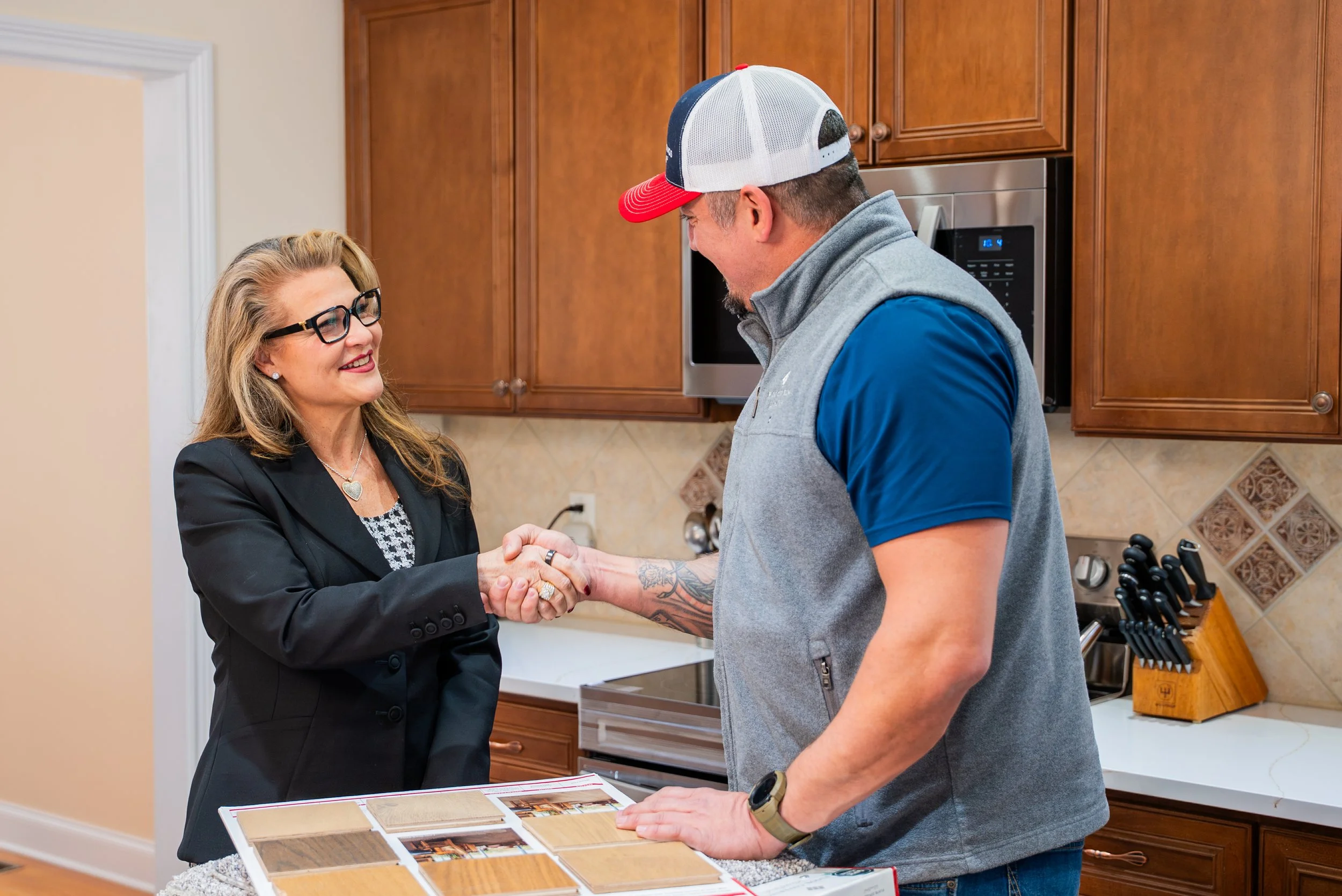 A woman and a man shake hands in a kitchen while looking at samples of wood flooring or countertops on a display board. The woman wears glasses and a black blazer, and the man has a baseball cap and casual clothing.