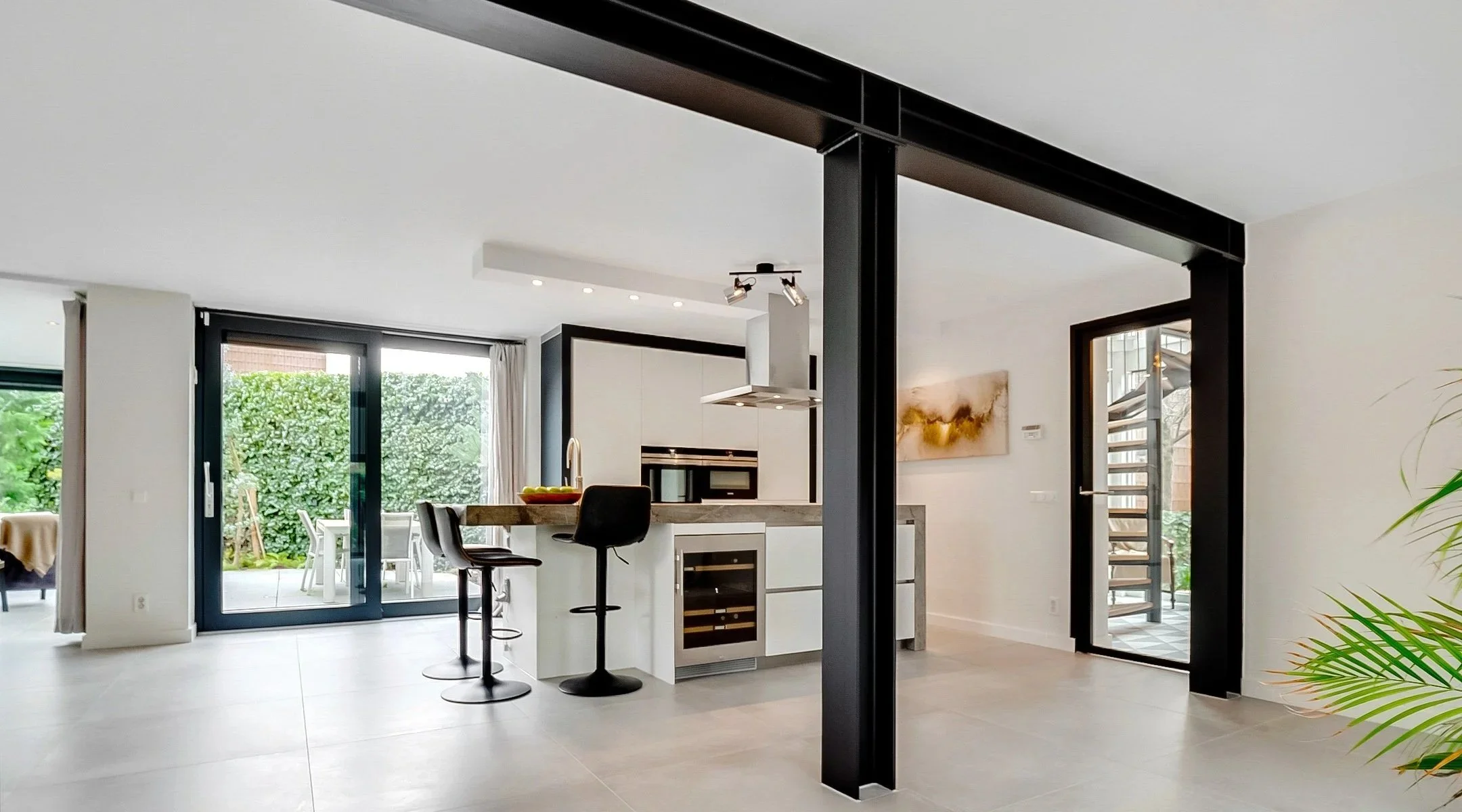 Moderne open keuken en eetruimte met zwarte stalen balken, zitplaatsen aan het kookeiland en openslaande deuren naar de tuin.