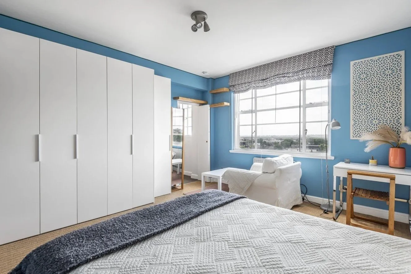 Blauwe slaapkamer met witte kasten, een bureau en een groot raam