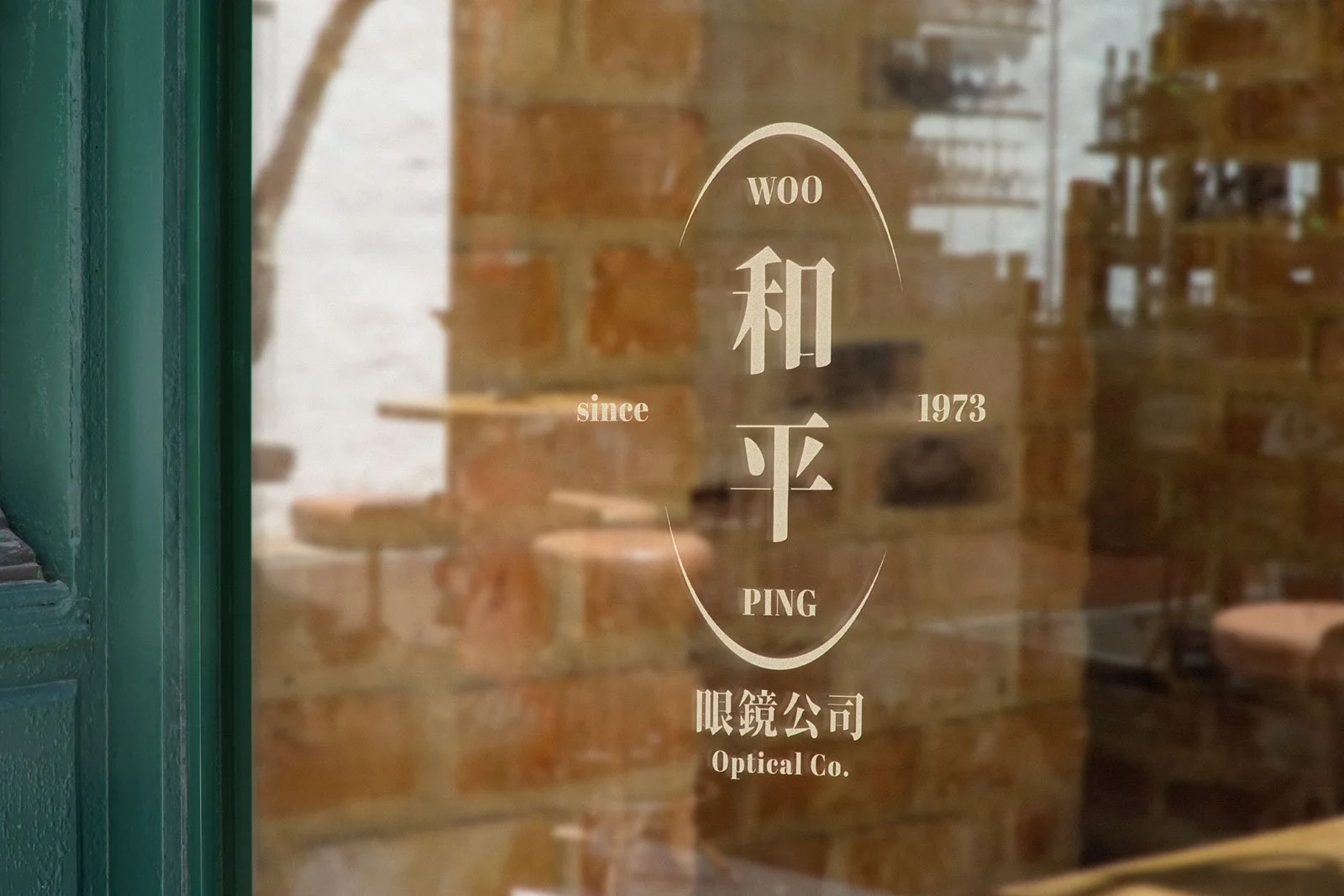 Glass door with store logo for WOO HEH PING Optical Co., established in 1973, with reflections of interior seating and brick walls.