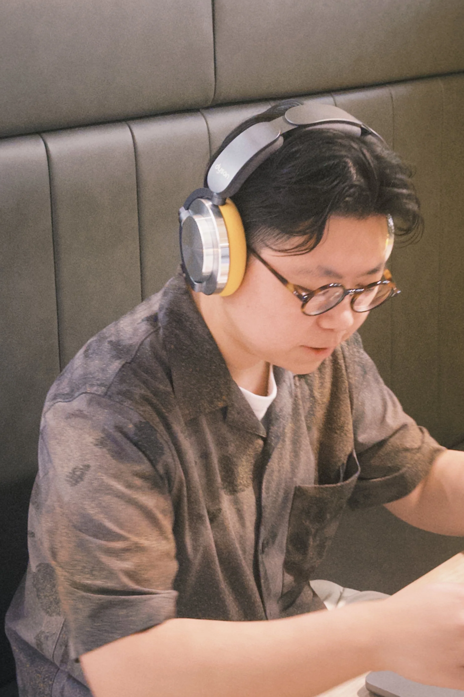 A young man with black hair, glasses, wearing a brown shirt, listening to music with large over-ear headphones, sitting at a table in a restaurant or cafe.