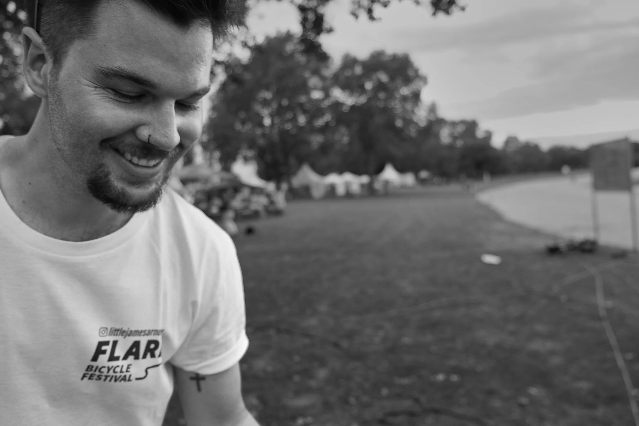 Ein junger Mann mit dunklem Haar und Bart lächelt bei einem Fahrrad-Festival im Freien, umgeben von Bäumen und Zelten.