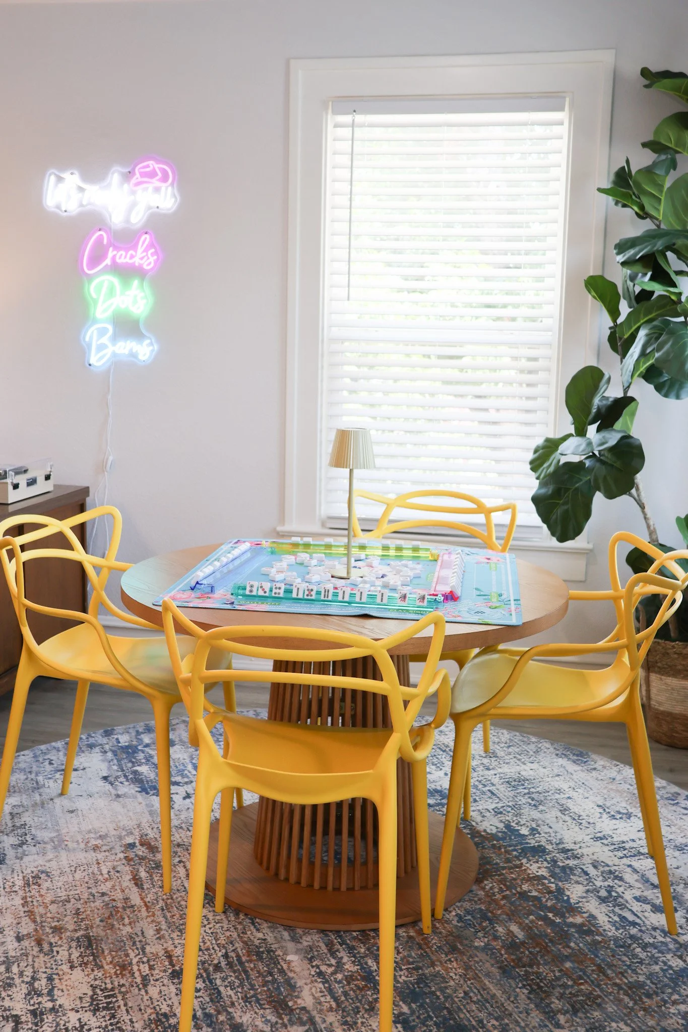 A round wooden table with a colorful Monopoly game set on it, surrounded by eight yellow chairs with curved backs. There is a large window with white blinds, a lamp with a pleated shade, a large green plant, and a neon sign on the wall that says 'Cra