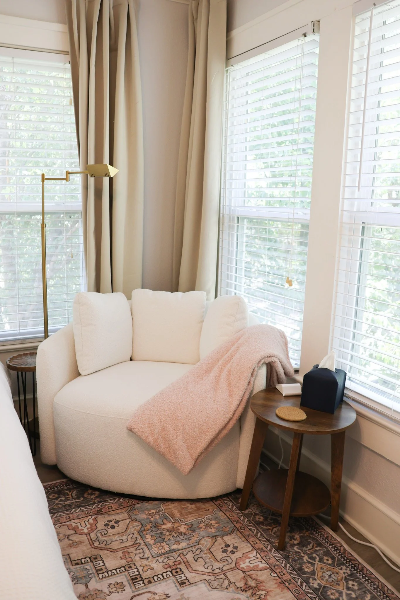 Living room with a cream-colored armchair, a pink throw blanket, a wooden side table, white window blinds, cream-colored curtains, a tall floor lamp, a patterned rug, and a small speaker.