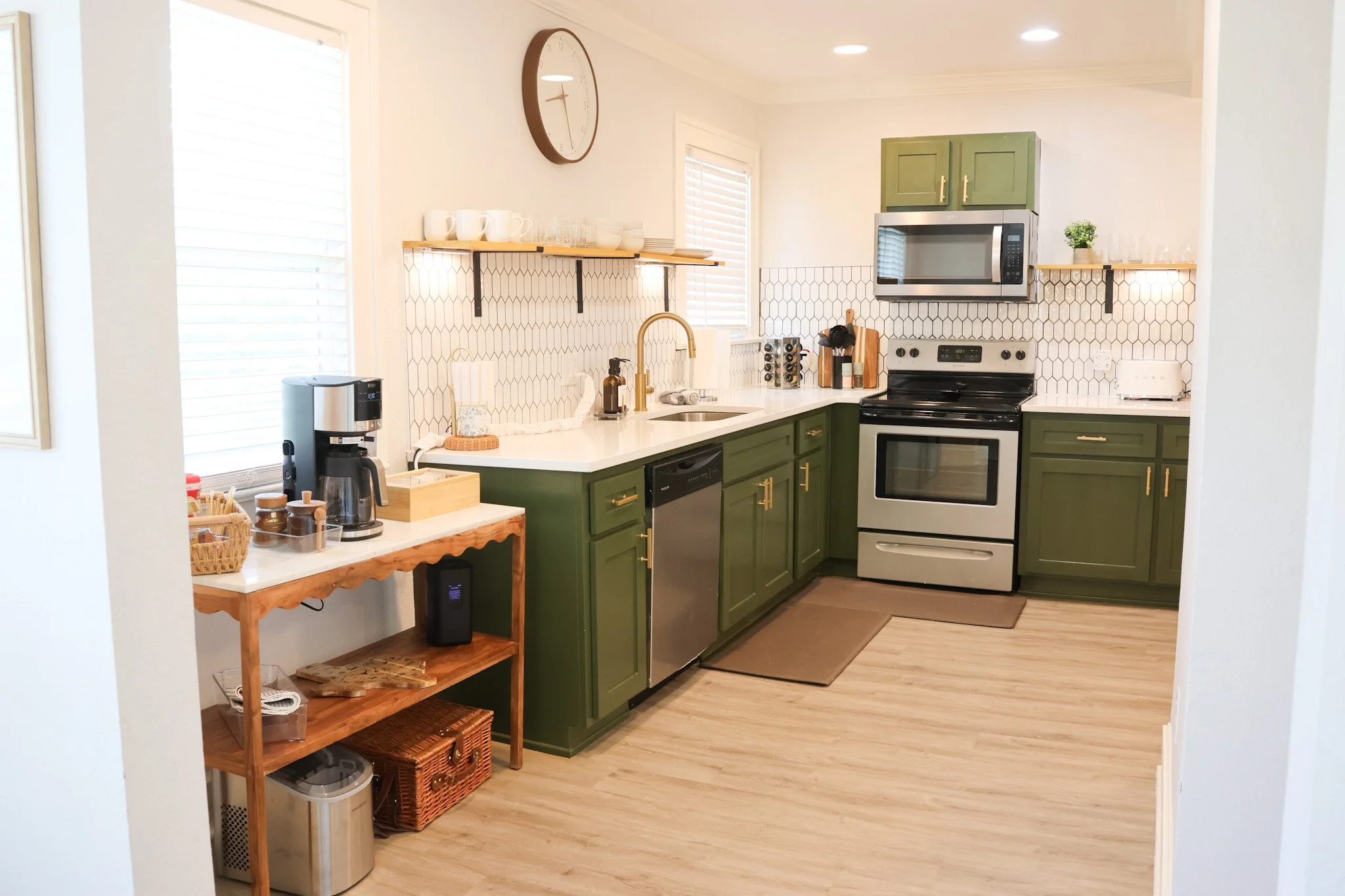 A modern kitchen with green cabinets, white countertops, a stainless steel oven, microwave, and a coffee machine on a wooden table. There are open shelves with dishes and cups, a wall clock, and two windows with blinds.