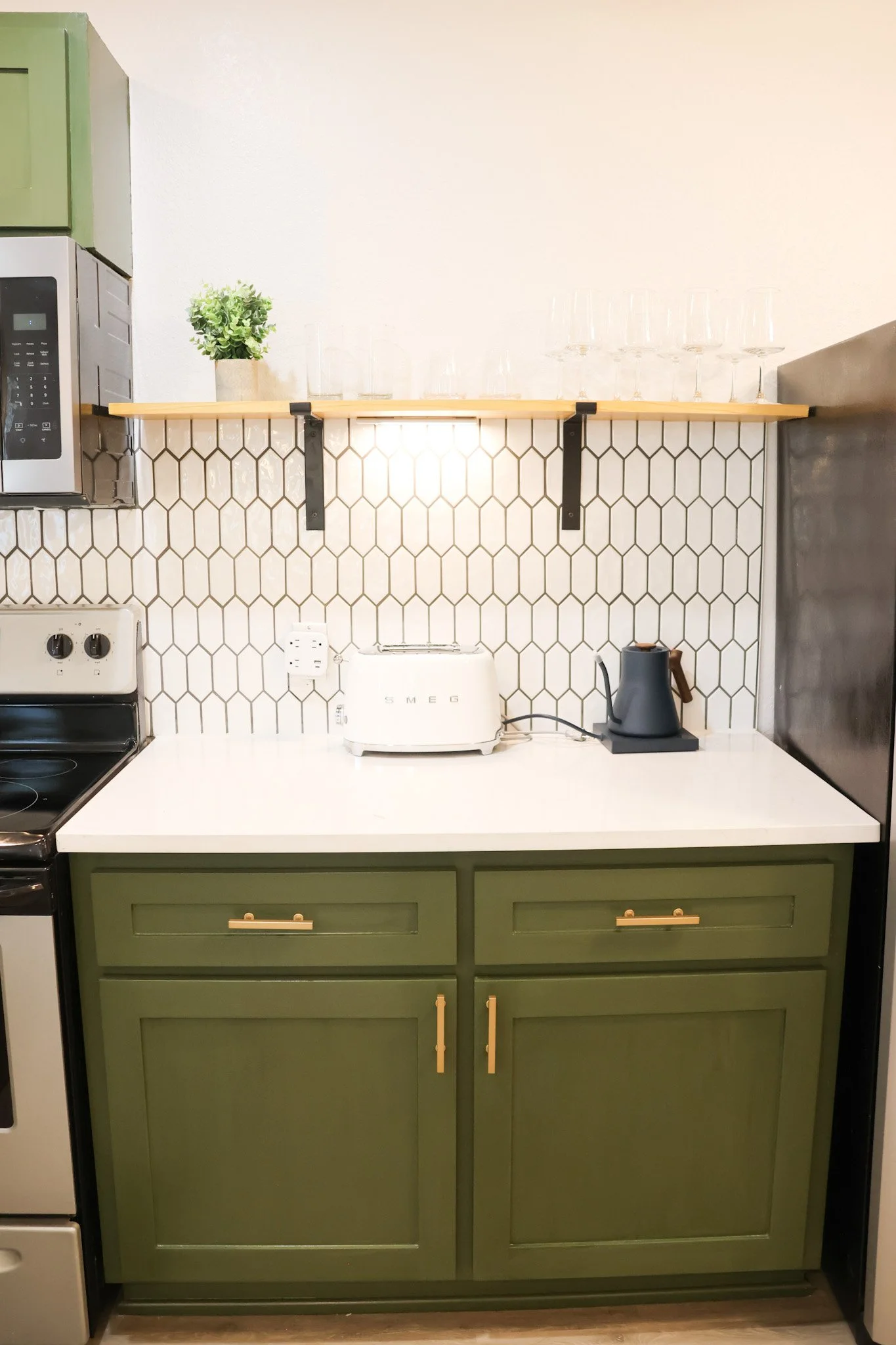 Kitchen with green cabinets, white countertop, honeycomb white tile backsplash, wooden floating shelf holding a potted plant and glassware, appliances including microwave, toaster, and kettle.