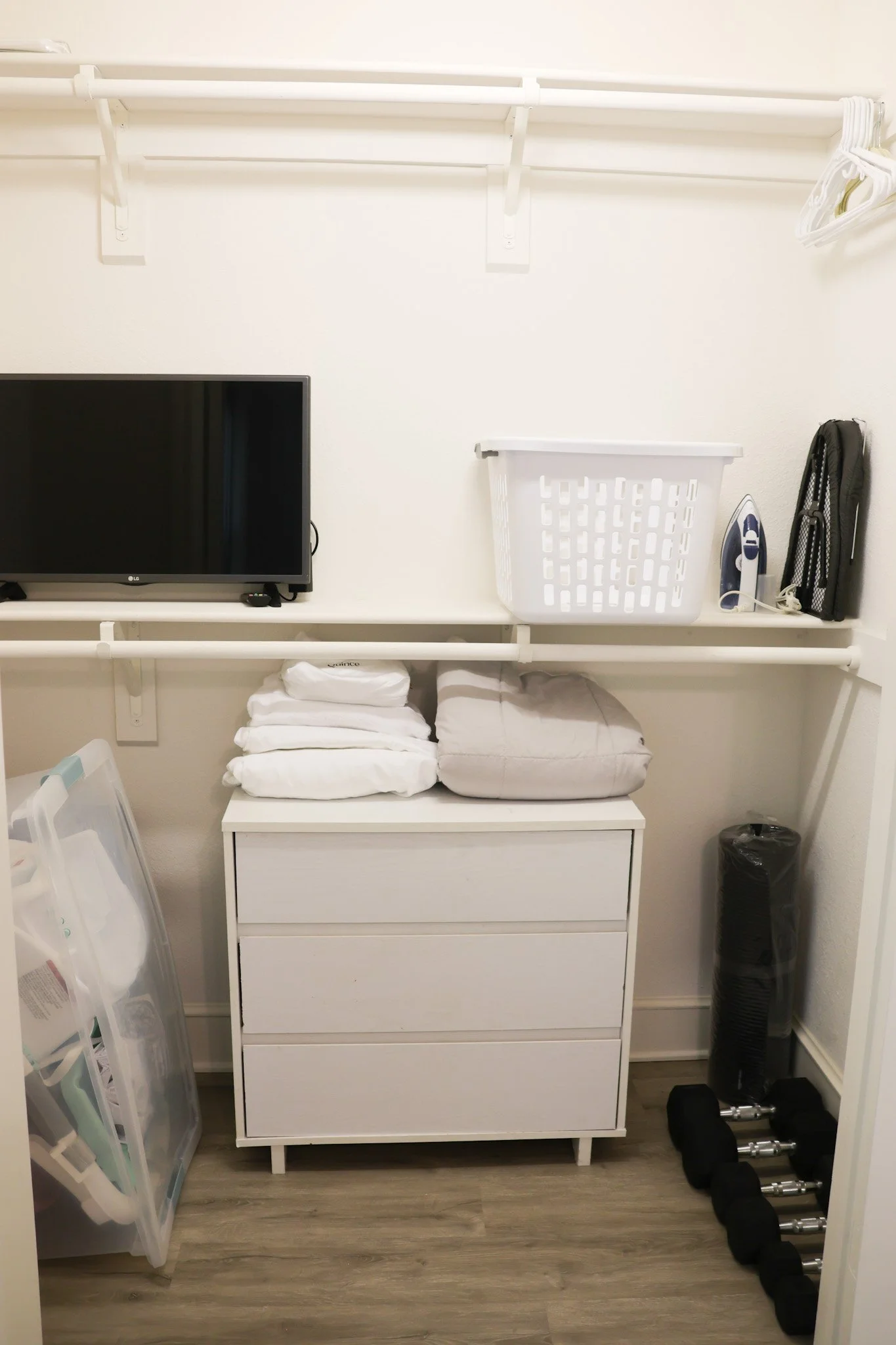 A closet with white shelving and a white dresser. On the shelves, there is a black television, a white laundry basket, an iron, and a black organizer with hangers. On the lower shelf, there are neatly folded white towels. To the right of the dresser,