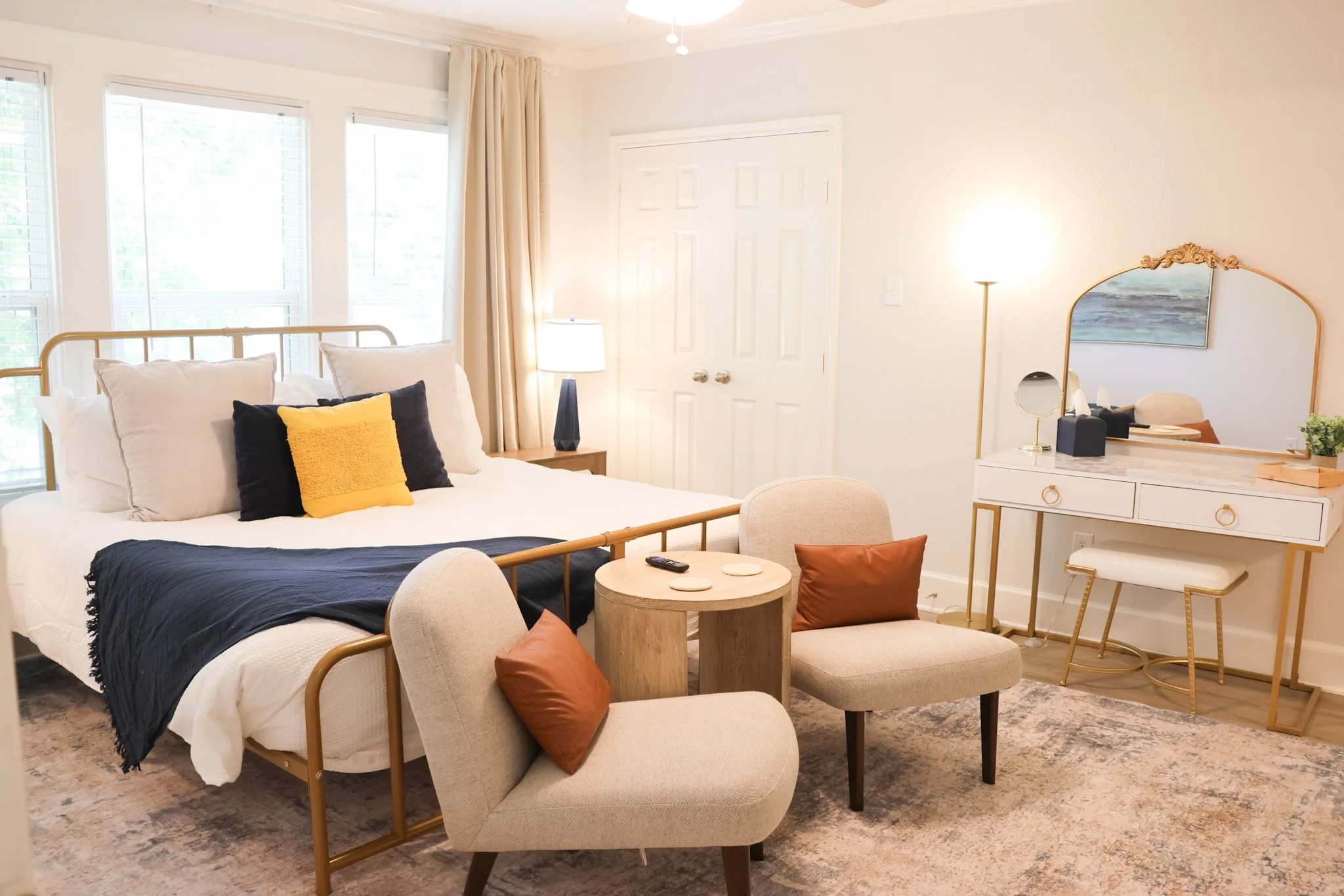 Bright bedroom with large windows covered by white blinds, a bed with white bedding and black, yellow, and beige pillows, a small round coffee table, an upholstered armchair with rust-colored pillows, a vanity with a mirror, black tissue box, and potted plant, and soft lighting.