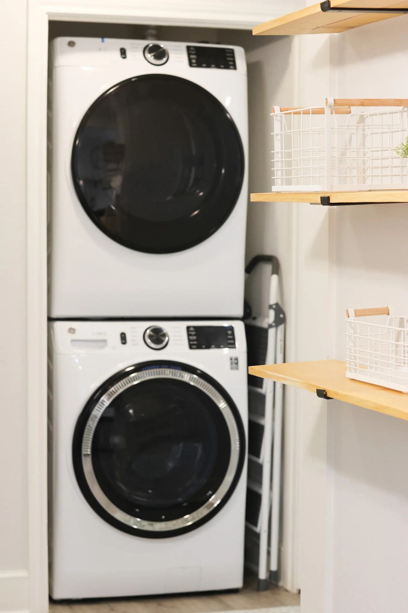 A laundry area with a stacked front-loading washer and dryer in an enclosed space. To the right are two wooden shelves with wire baskets. A small potted plant is on the top shelf.