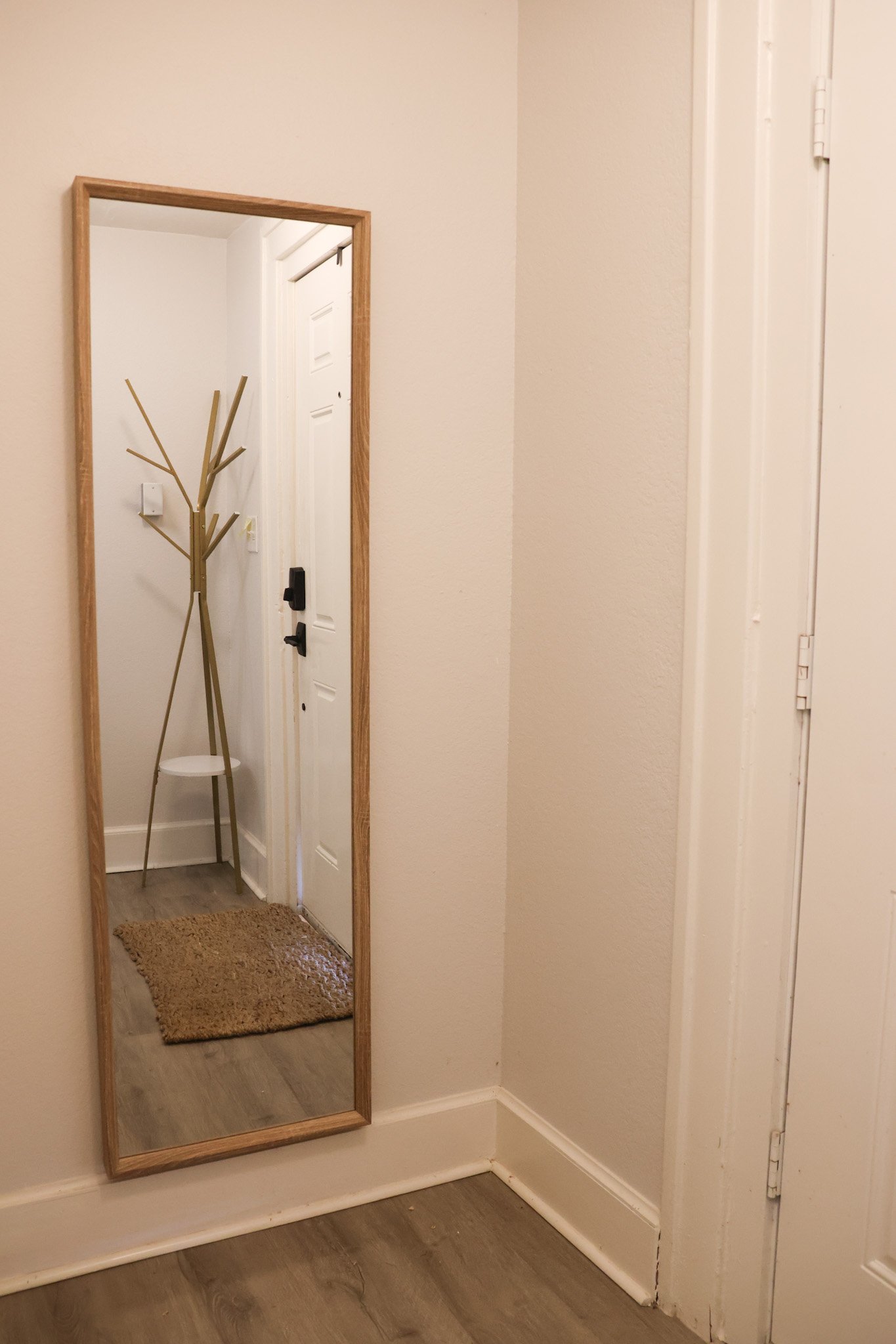 A large rectangular mirror on a wall showing the image of a hallway entrance with a coat rack and a door, standing on wooden flooring with a small brown rug in front.