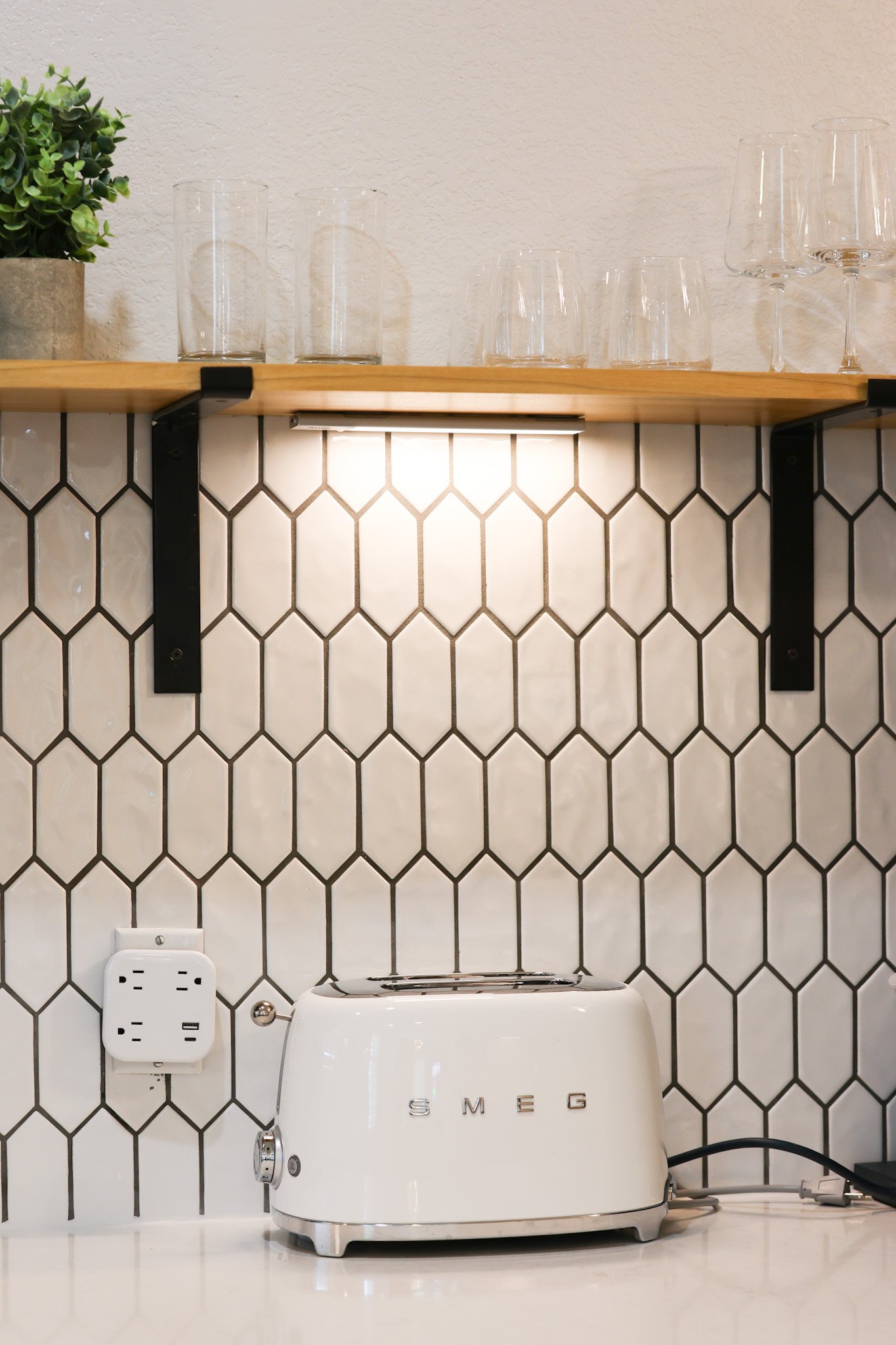 A white SMEG toaster on a kitchen countertop below a wooden shelf with glasses and a plant, against a white textured wall with black hexagonal tile backsplash.
