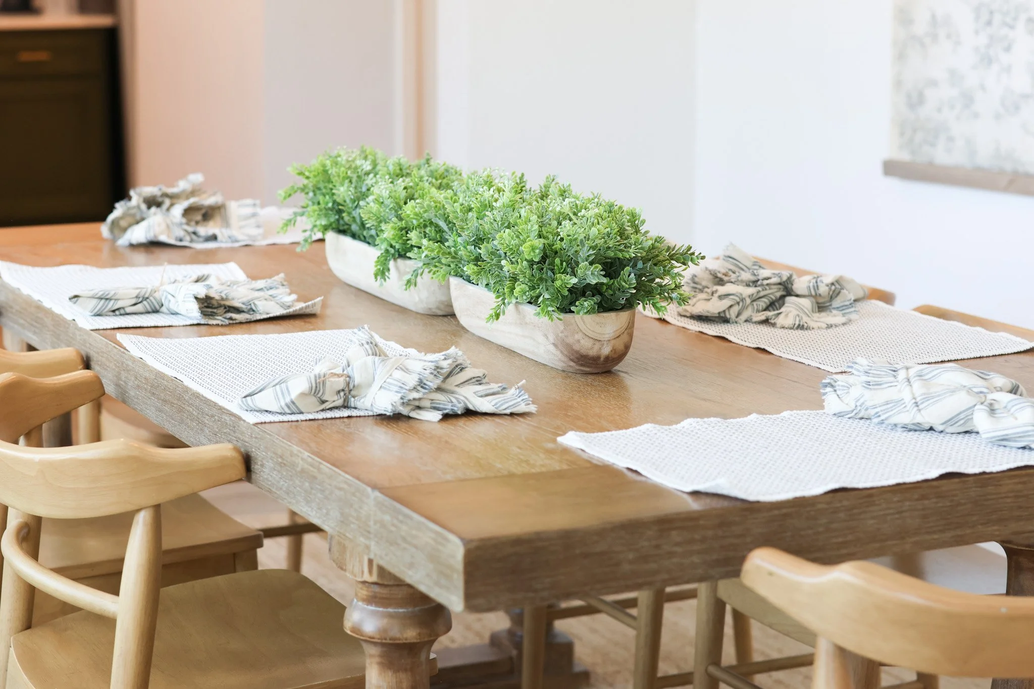 A wooden dining table decorated with green potted plants and patterned cloth napkins, surrounded by wooden chairs in a room with white walls and a partial view of a window.