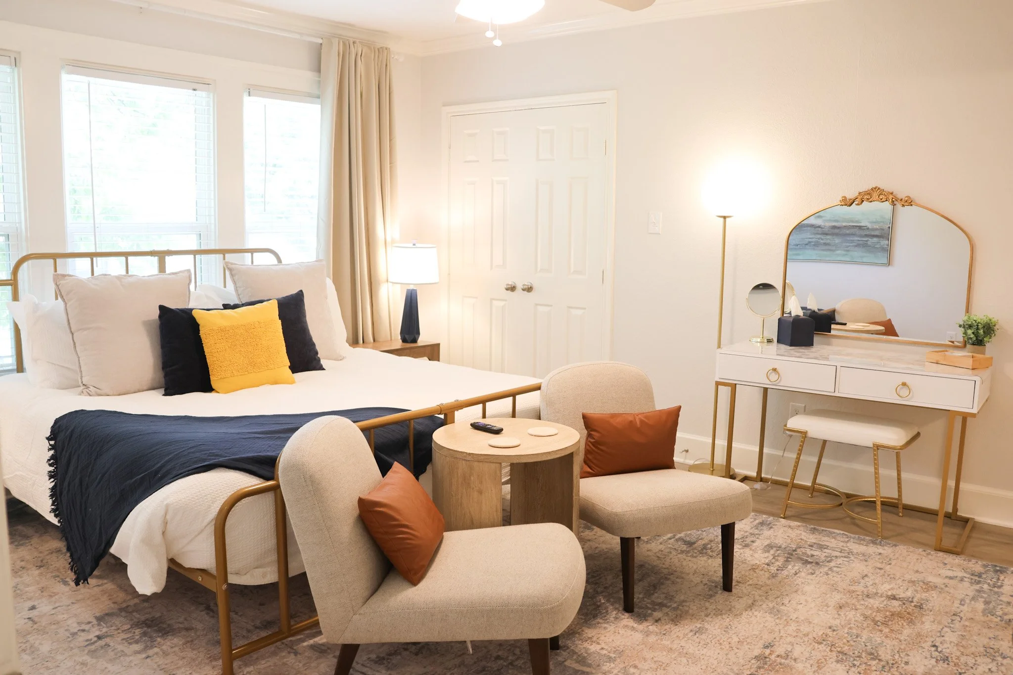 A cozy bedroom with a neatly made bed covered in white bedding, layered with cream, navy, and yellow pillows. Two large windows with white blinds and beige curtains let in natural light. Next to the bed, there is a wooden nightstand with a modern bla
