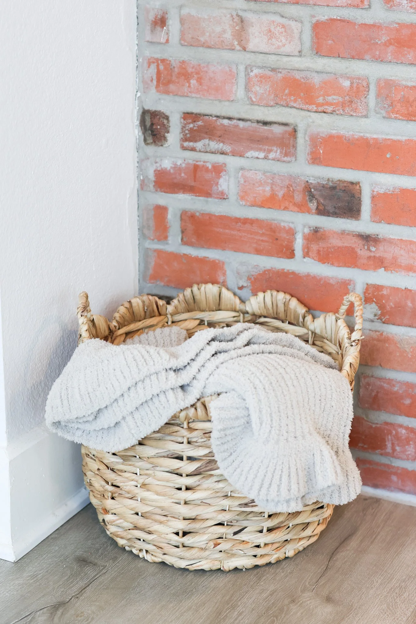 A woven basket with a plush, cream-colored blanket inside, placed on a wooden floor against a background of a white wall and a red brick wall.