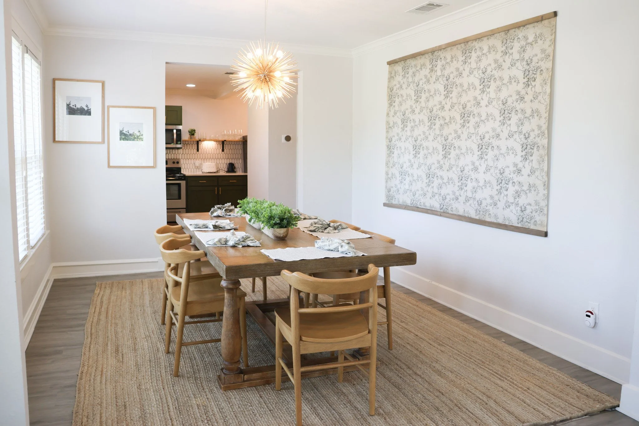 A dining room with a wooden table set with six placemats, napkins, and a centerpiece, surrounded by beige chairs, a large floral artwork on the wall, and a modern chandelier overhead.