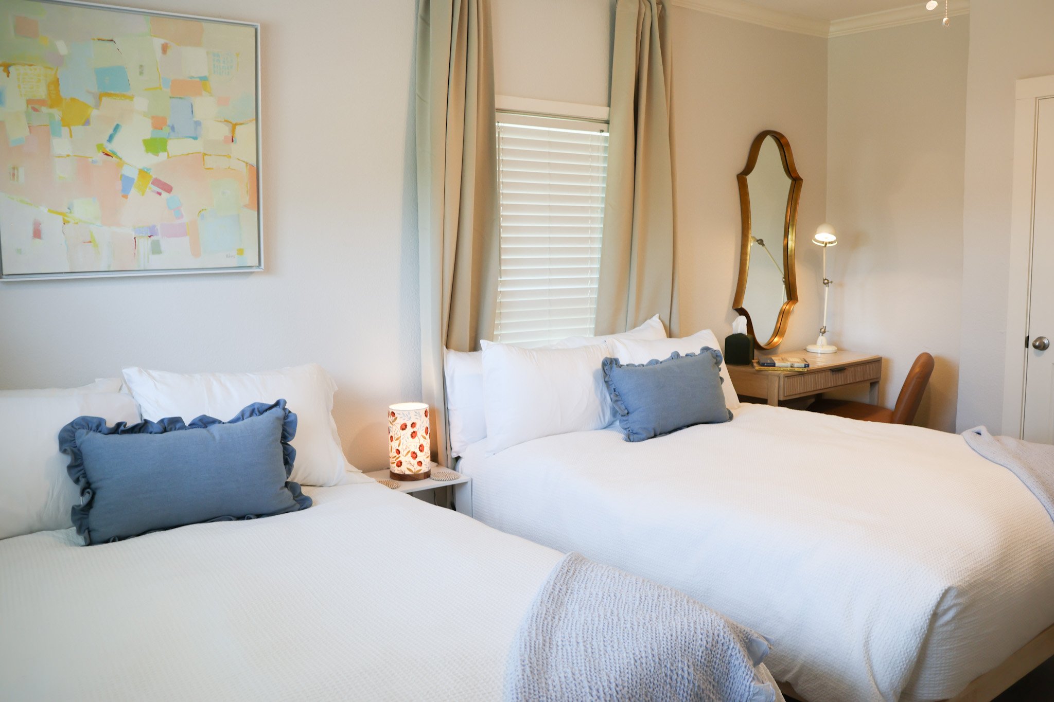 A cozy bedroom features two neatly made beds with white linens and blue ruffled pillows. A small bedside table holds a decorative lamp with a floral pattern. The wall above the beds has a large abstract painting with pastel colors. A window with clos