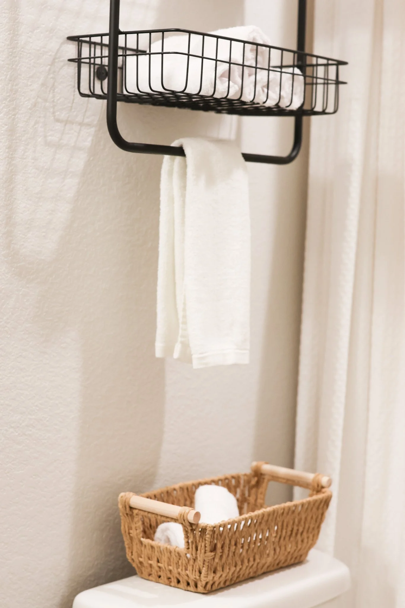 Bathroom storage with a black metal wall-mounted basket containing rolled white towels, a small woven basket with rolled towels inside, mounted on a beige wall near a window with white curtains.