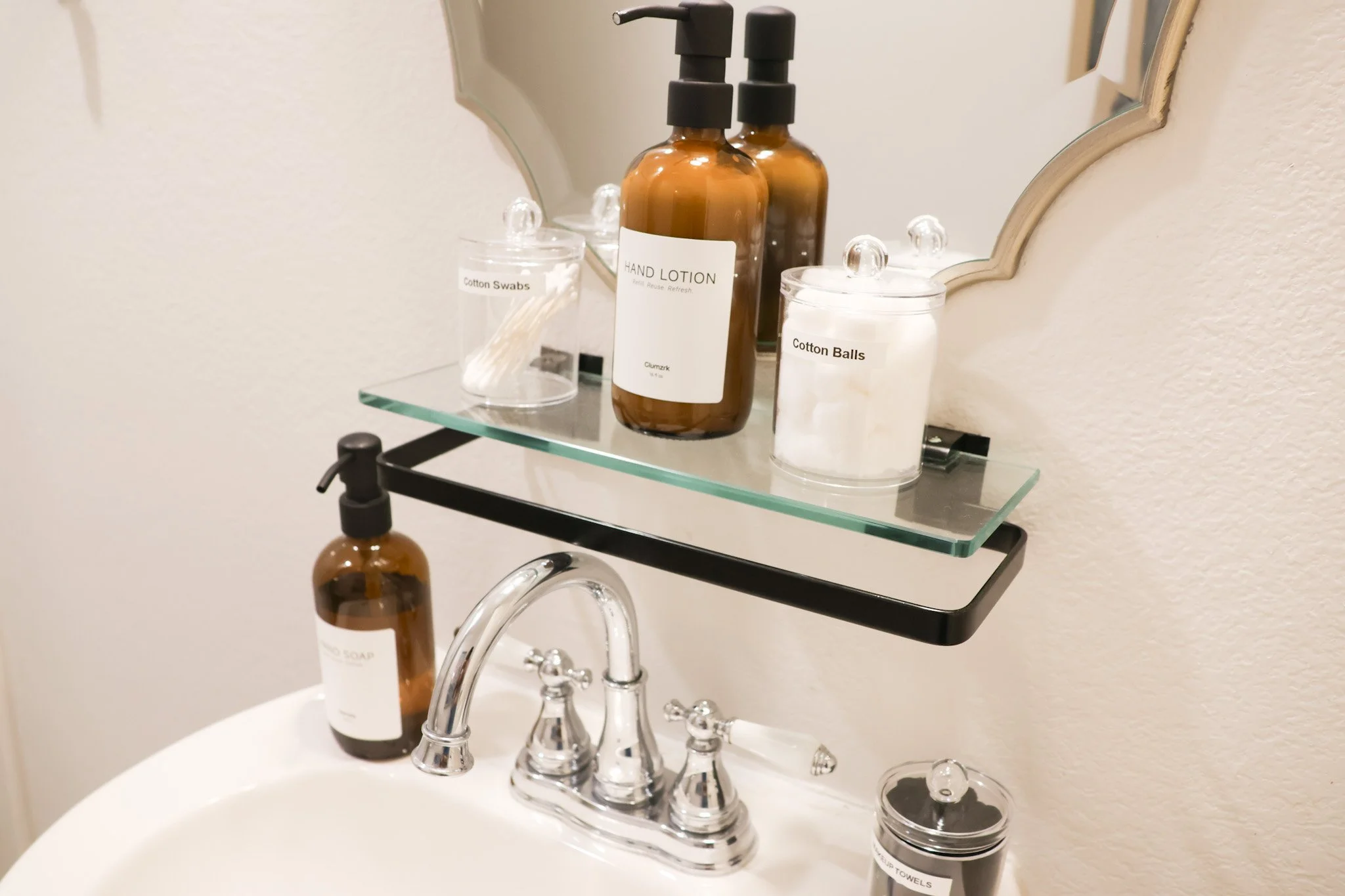 Bathroom sink with a mirror, holding bottles of hand lotion and soap, cotton swabs, cotton balls, and a jar of cotton pads on a glass shelf.