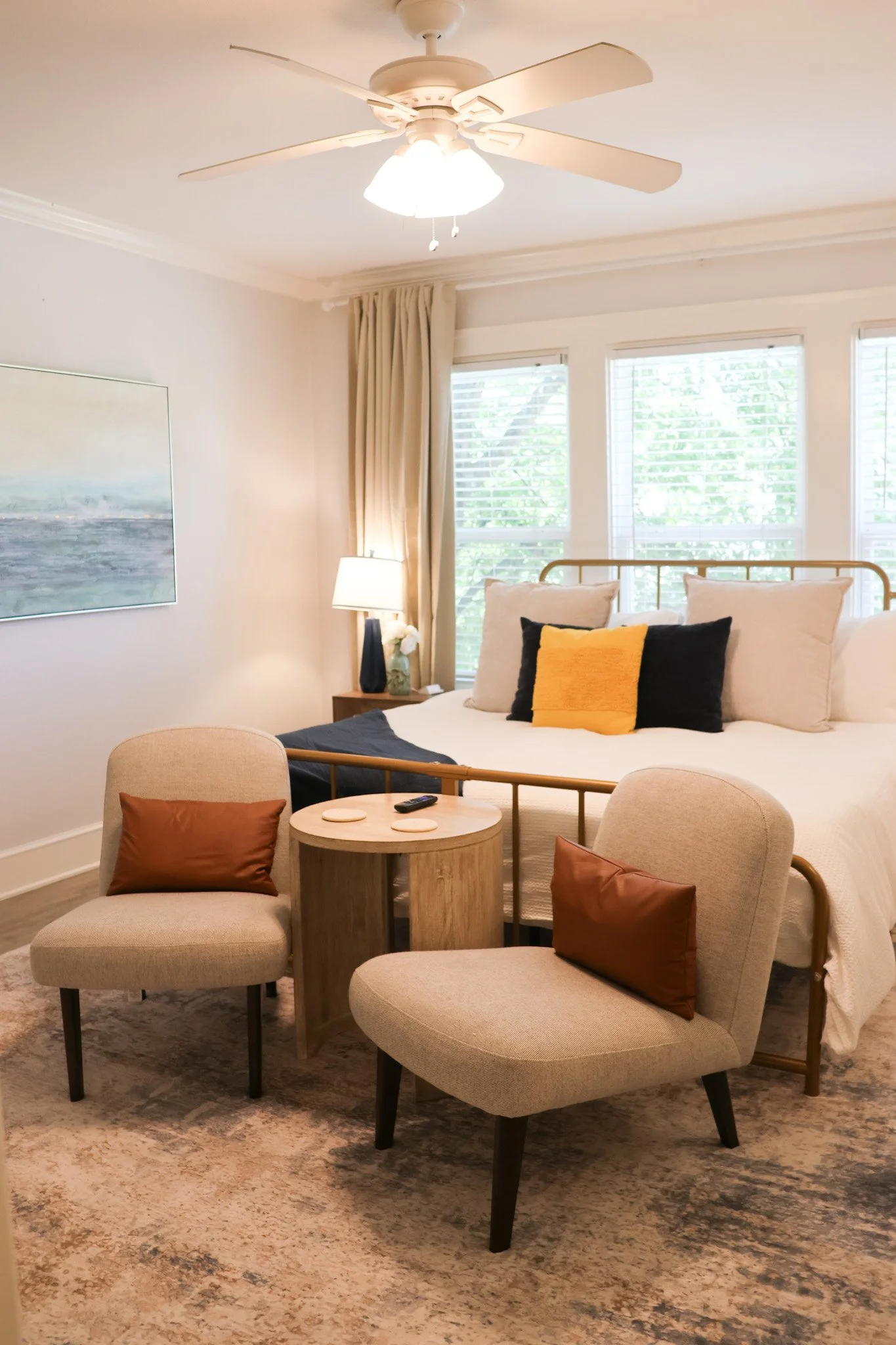 A bedroom with a bed, pillows, bedside table with a lamp, windows with white blinds, a ceiling fan, and a sitting area with two beige chairs and a round wooden table.