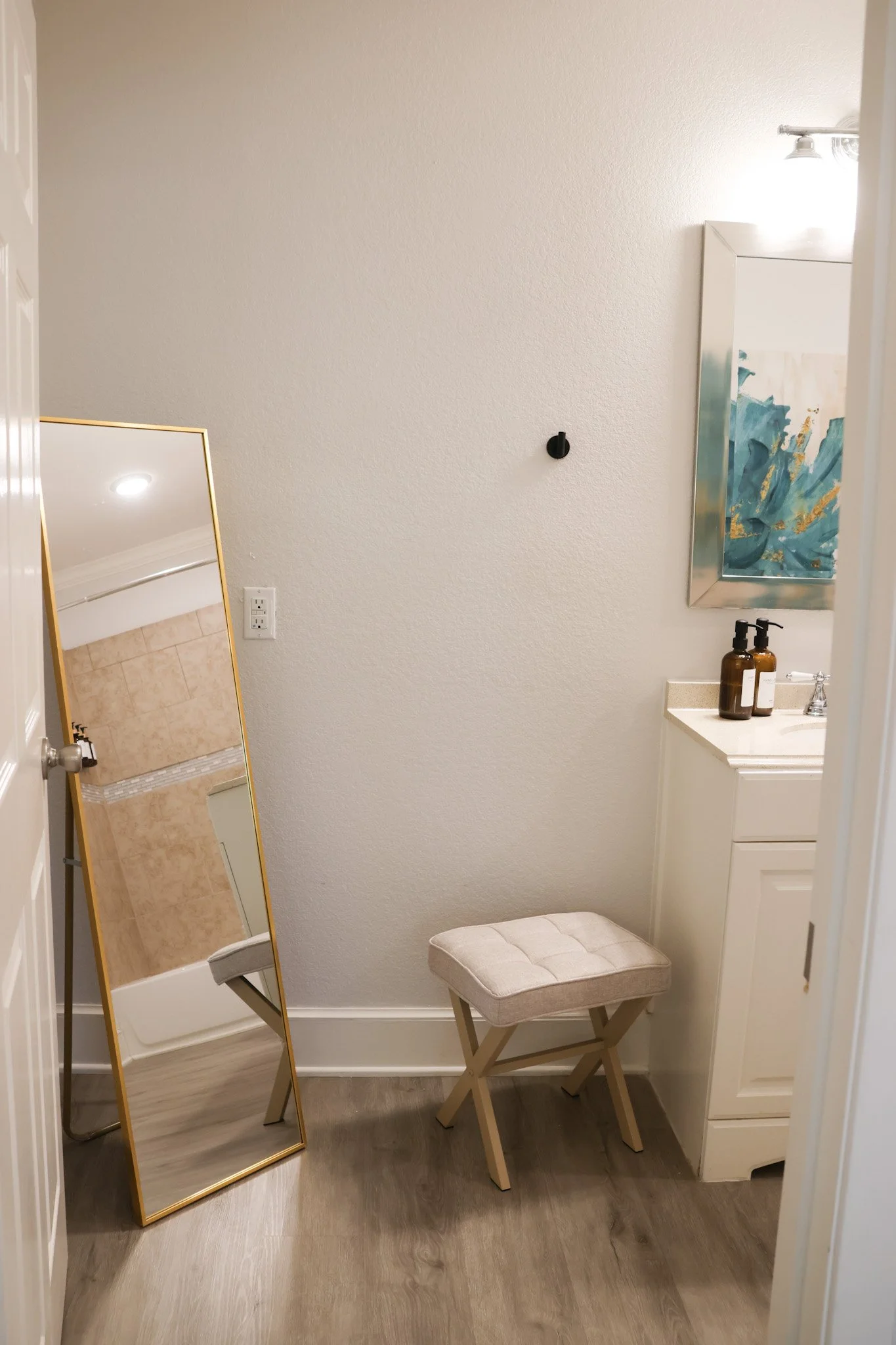 A small bathroom corner with a full-length mirror with a gold frame, a beige padded stool, a white cabinet, and a small vanity with two amber-colored bottles. There is a white wall with a light fixture and an abstract blue and gold framed artwork.
