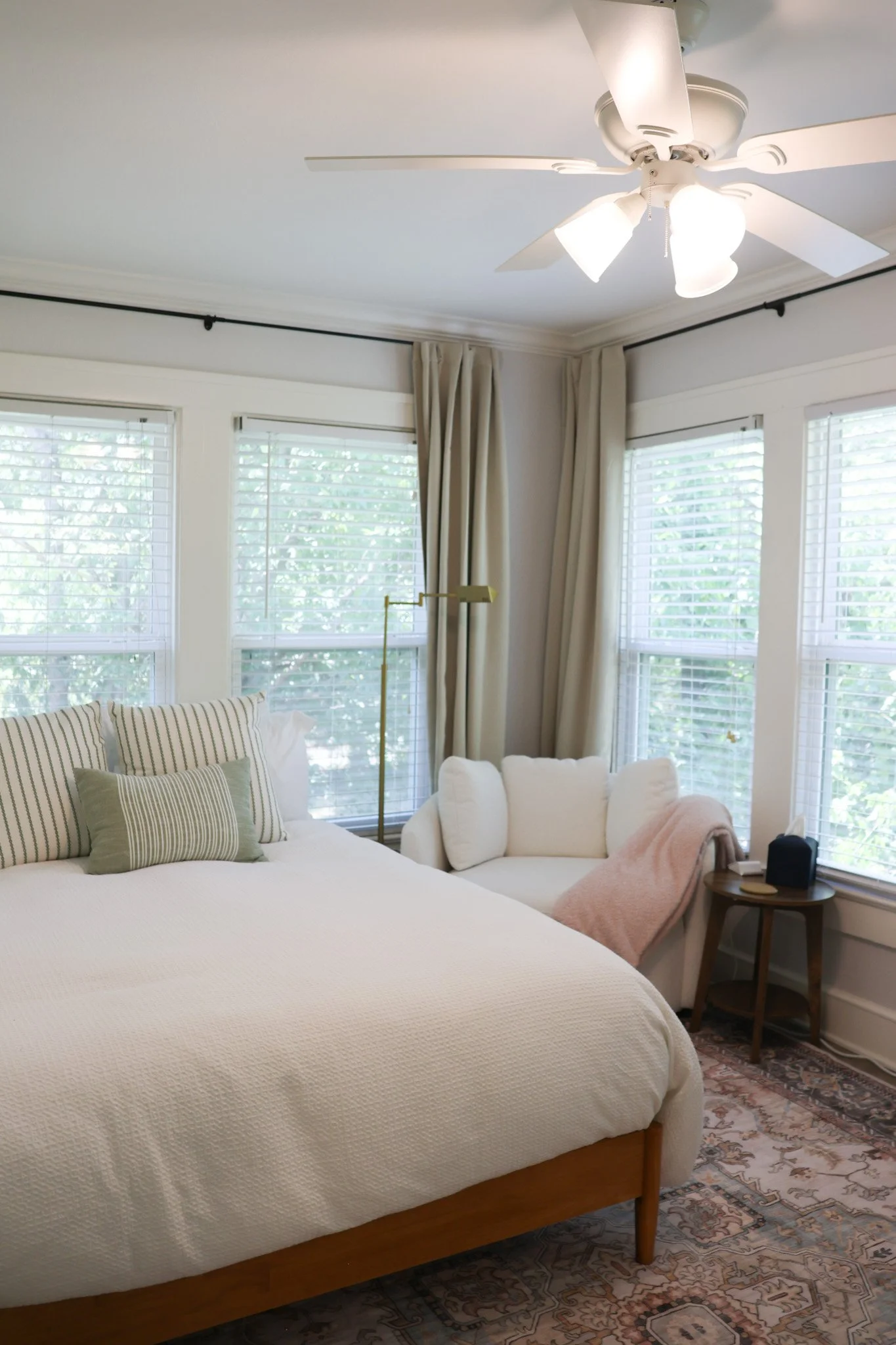 A bedroom with a large white bed, striped pillows, a small side table with a black alarm clock, beige curtains, and large windows with white blinds. A ceiling fan with lights is turned on.