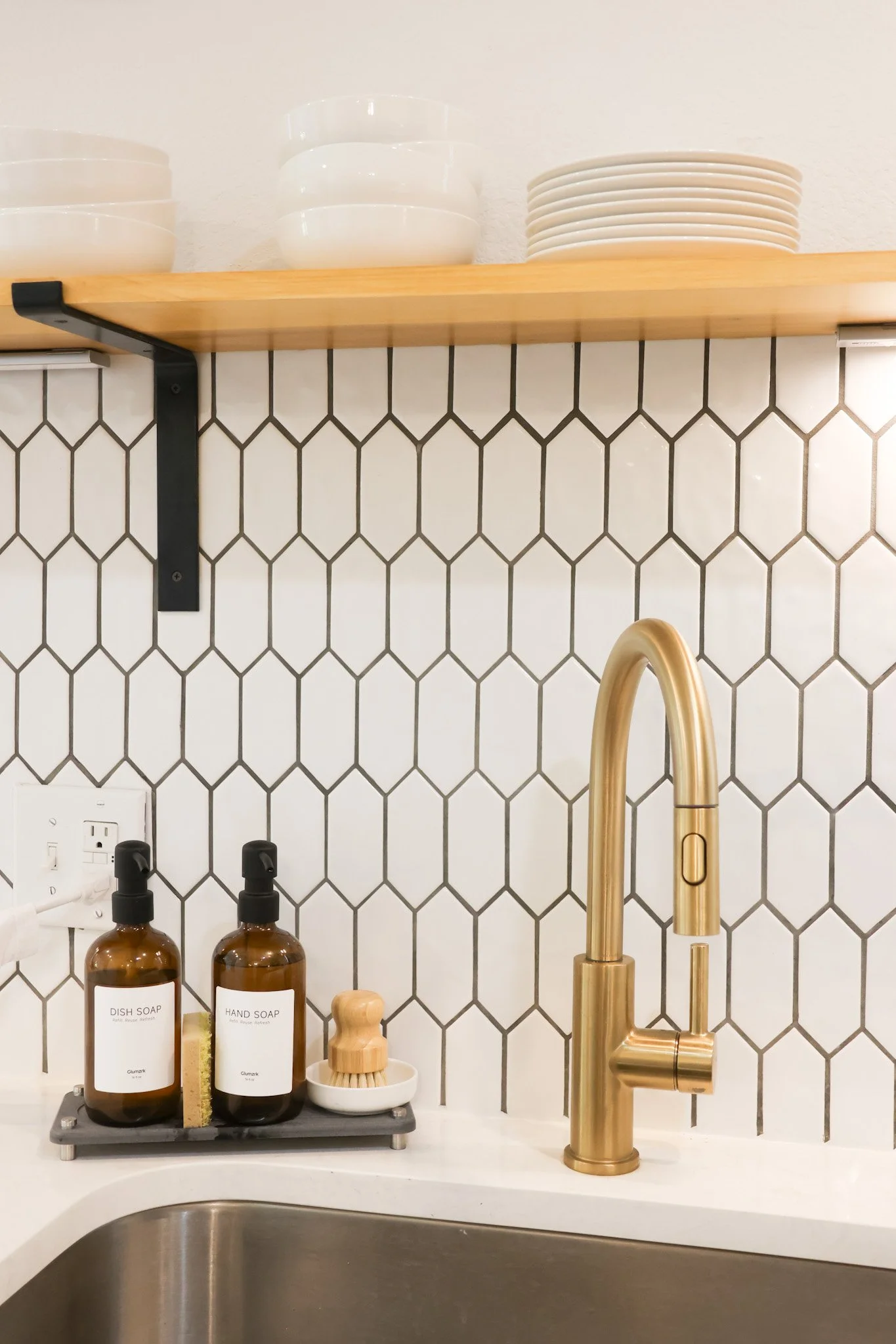 A kitchen countertop with brown soap dispensers, a wooden scrub brush, and a white soap dish, above a stainless steel sink, with a gold faucet and black and white hexagonal tile backsplash.