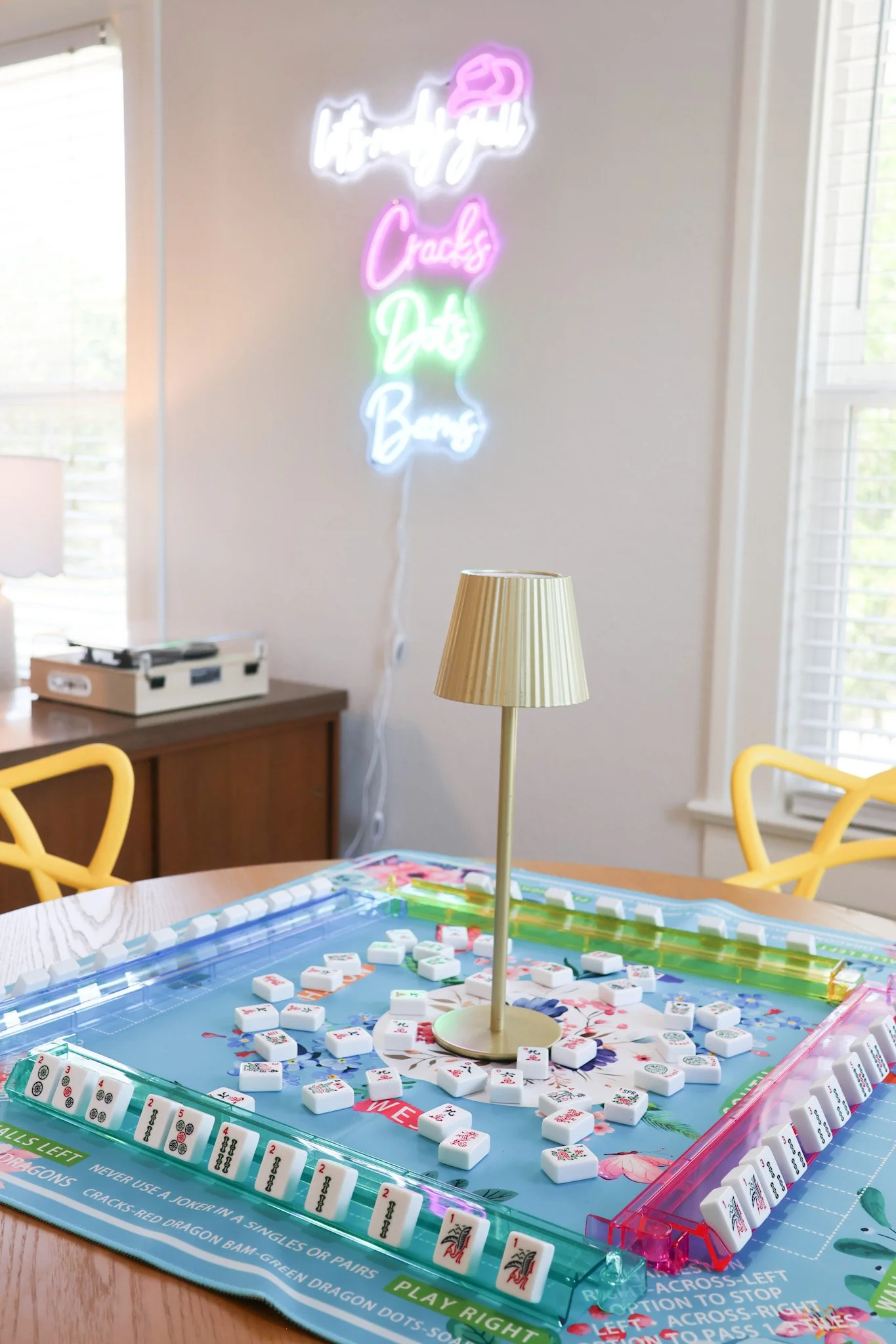 Close-up of a Mahjong game in progress on a wooden table, with Mahjong tiles laid out on a colorful game mat featuring floral designs and instructions. A gold table lamp is in the background, and a neon sign on the wall reads 'happy go Cracks, Dares,
