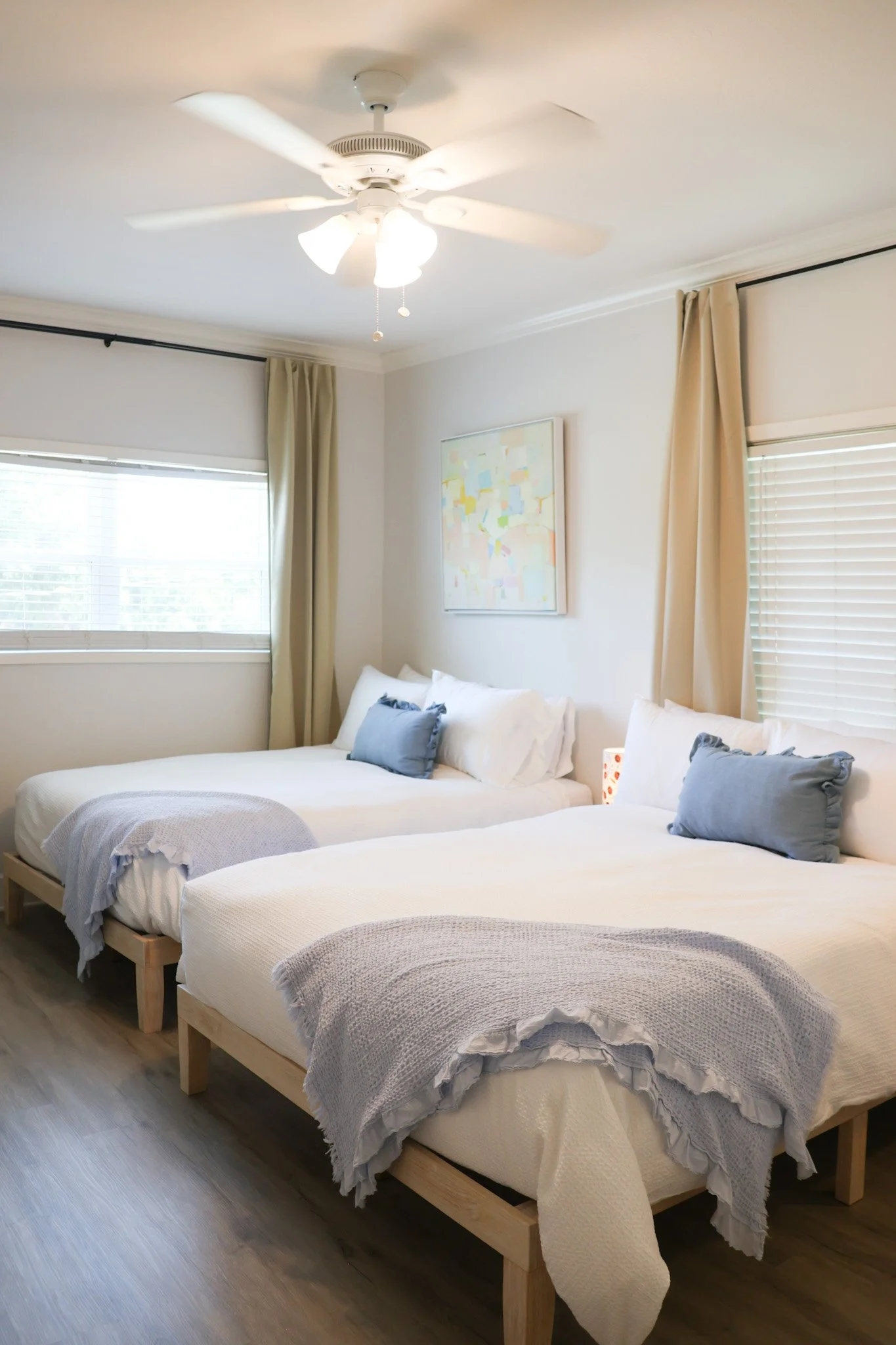 A bedroom with two beds having white bedding and blue pillows, a window with blinds and curtains, a ceiling fan with lights, and a piece of colorful abstract artwork on the wall.