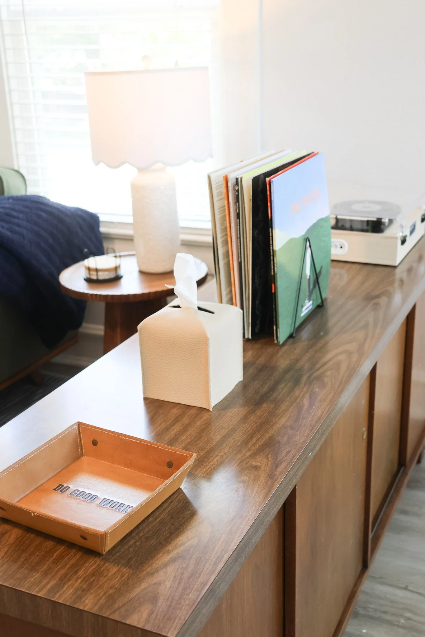 A wooden table with a tissue box, a tray with the text 'Do Good Work', a small tray, a paper holder, and a row of magazines or books standing upright. In the background, a lamp, a small speaker, and other objects are visible near a window with blinds