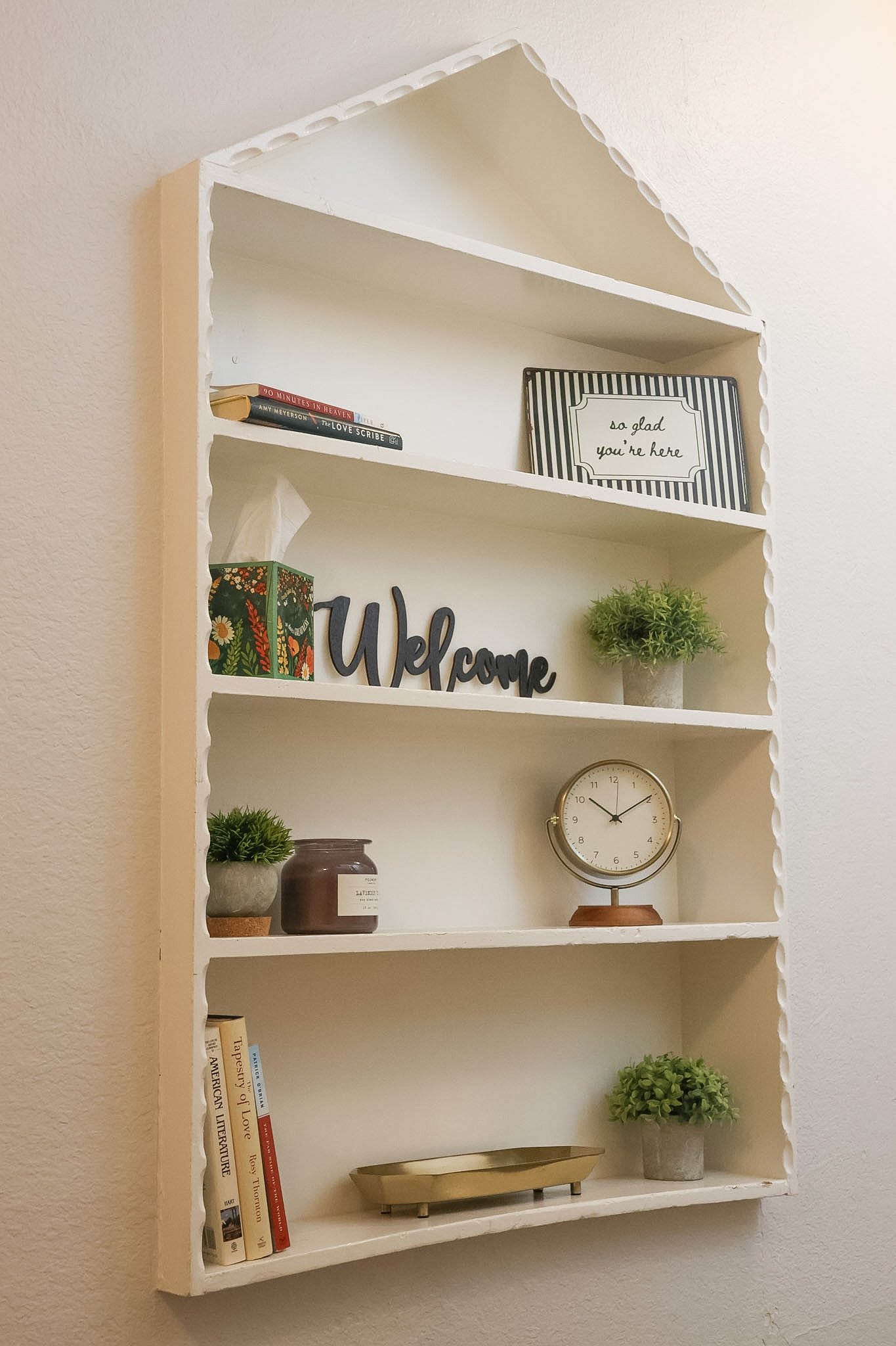 A white wall-mounted shelf with decorative scalloped edges, holding books, plants, a clock, and decorative signs.