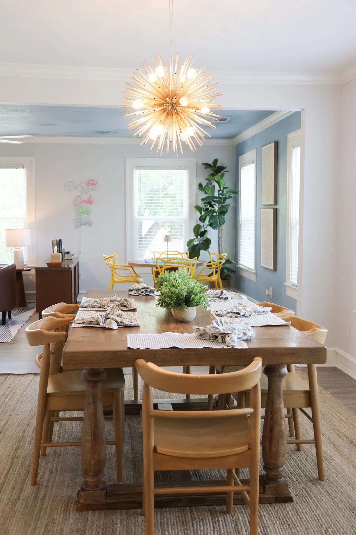 A dining room with a large wooden table set with striped napkins and a green plant centerpiece. Surrounding the table are mostly wooden chairs, some with armrests. The room features several windows with white blinds, a herringbone rug, a modern light