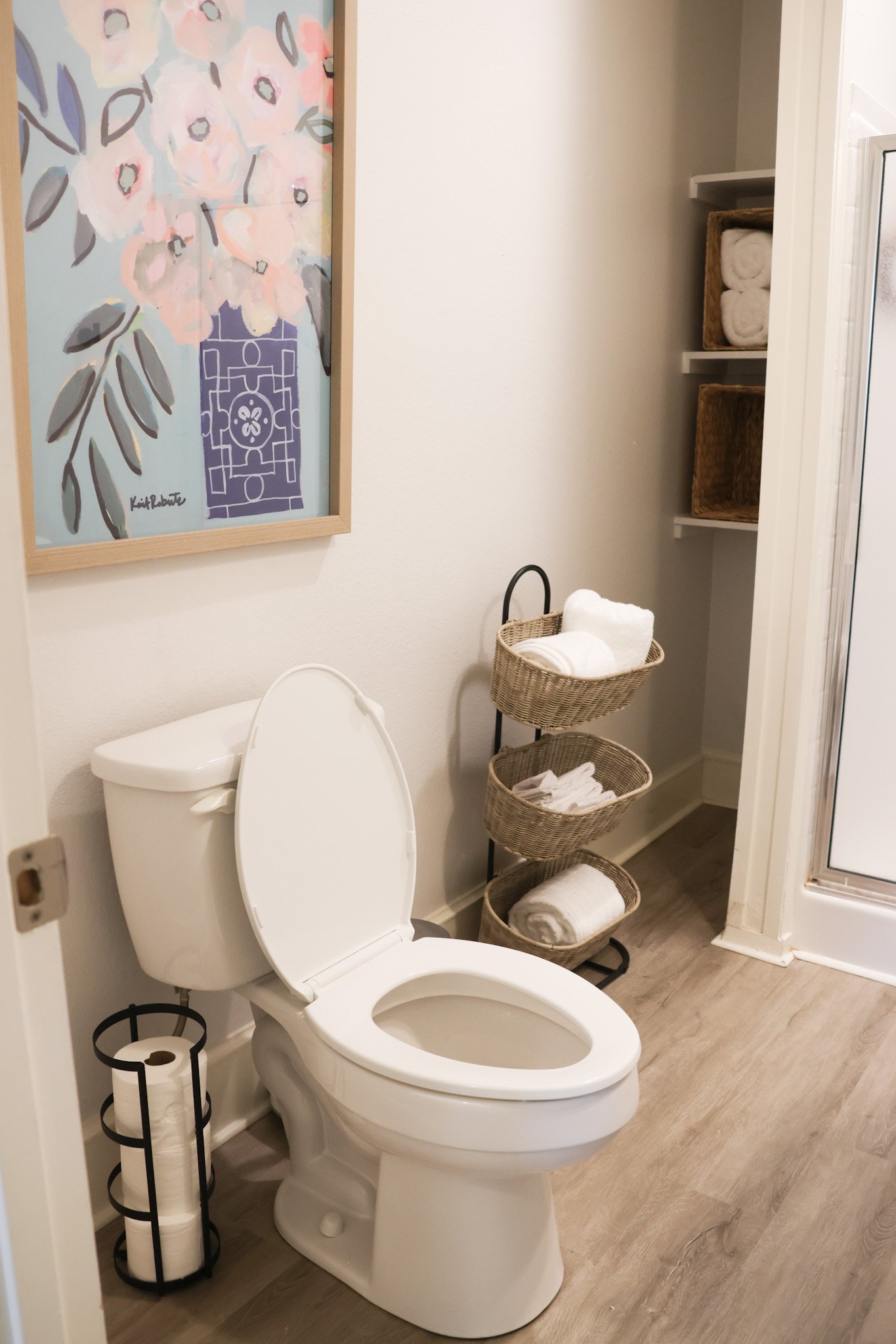 A bathroom with a white toilet, a black metal toilet paper holder, a three-tier woven basket with rolled towels and towels, a colorful abstract floral painting on the wall, and a white shelving unit with more rolled towels.