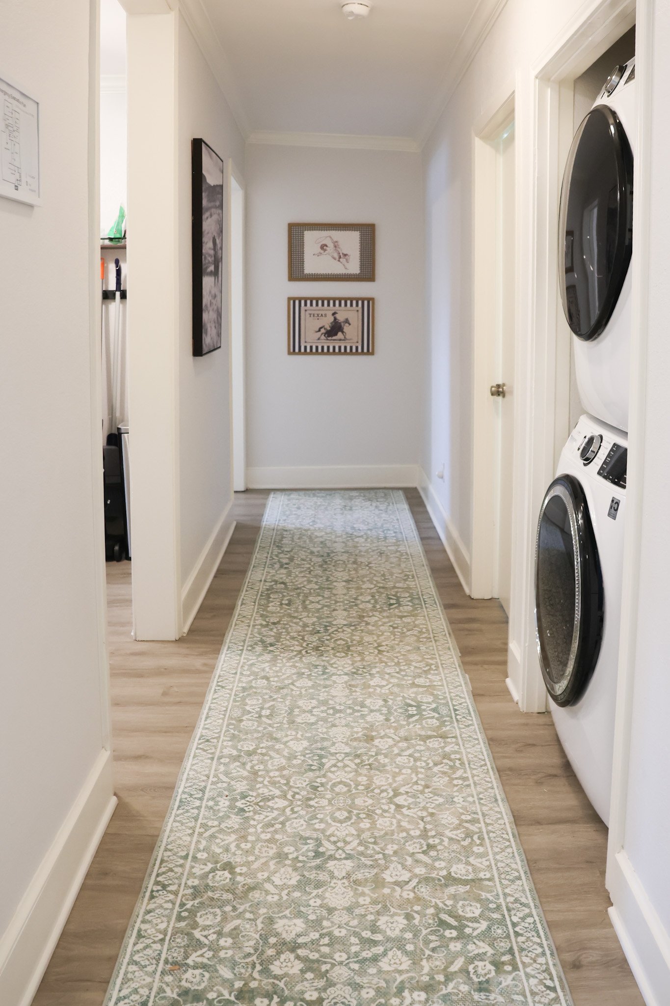 A hallway with light-colored walls and wooden flooring. There is a patterned runner rug on the floor. To the right, there is a stackable washer and dryer unit. The end of the hallway features two framed pictures on the wall, one of a horse and rider,