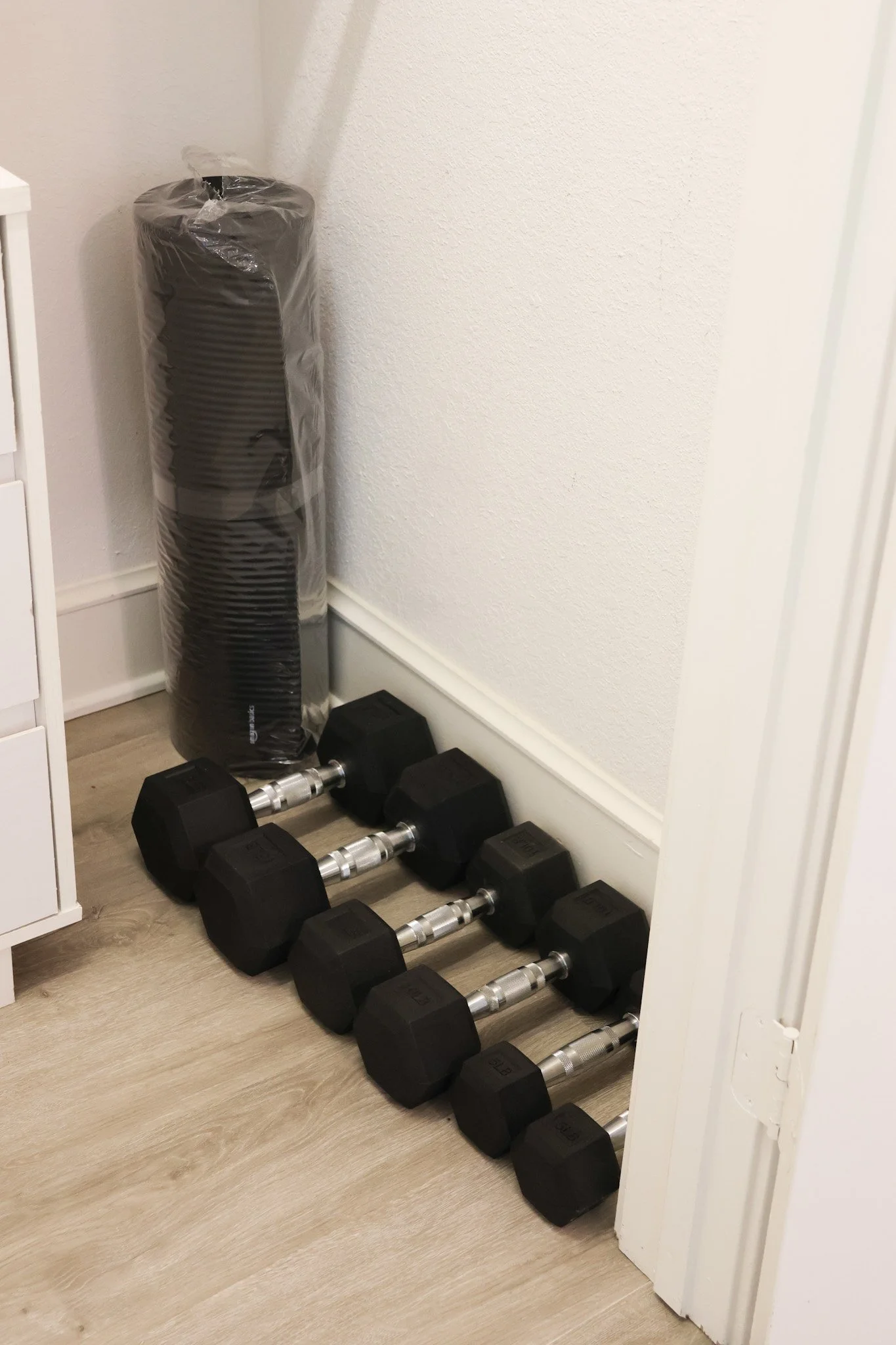 A set of black dumbbells with silver handles arranged on a light-colored wooden floor in front of a white wall, with a rolled-up yoga or exercise mat wrapped in plastic leaning against the wall behind them.