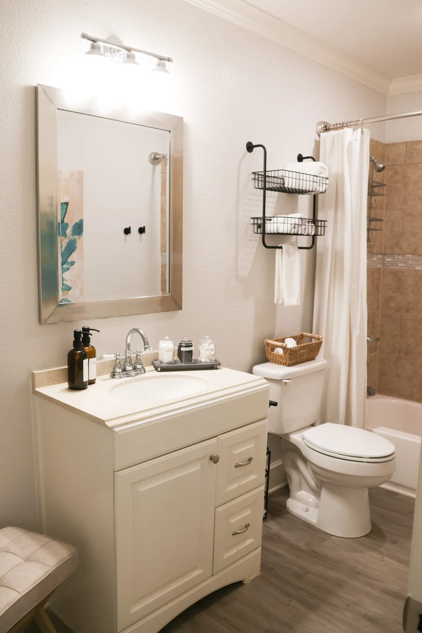 A bathroom with a white vanity, a mirror, a black wall-mounted towel rack with white towels, a toilet, and a shower with a curtain. The floor is wood-like tile.