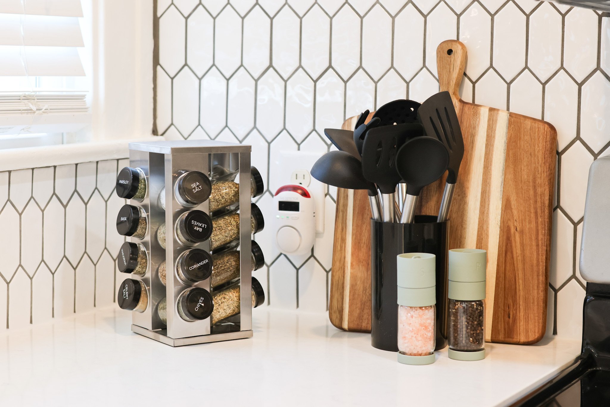 A kitchen countertop with a spice rack holding various spices, a wooden cutting board, and containers of pink Himalayan salt, pink pepper, and black peppercorns.