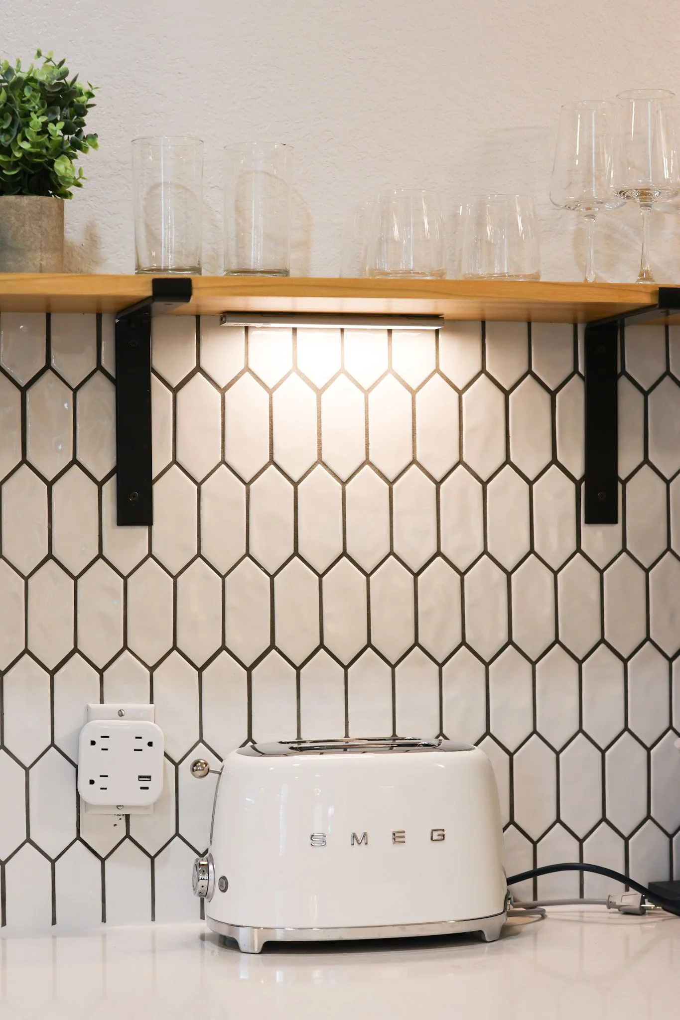 A white SMEG toaster on a white countertop with a black power cord plugged into a wall socket. Behind it, a black and white hexagonal tiled wall with a wooden shelf holding clear drinking glasses and a green plant in a concrete pot.