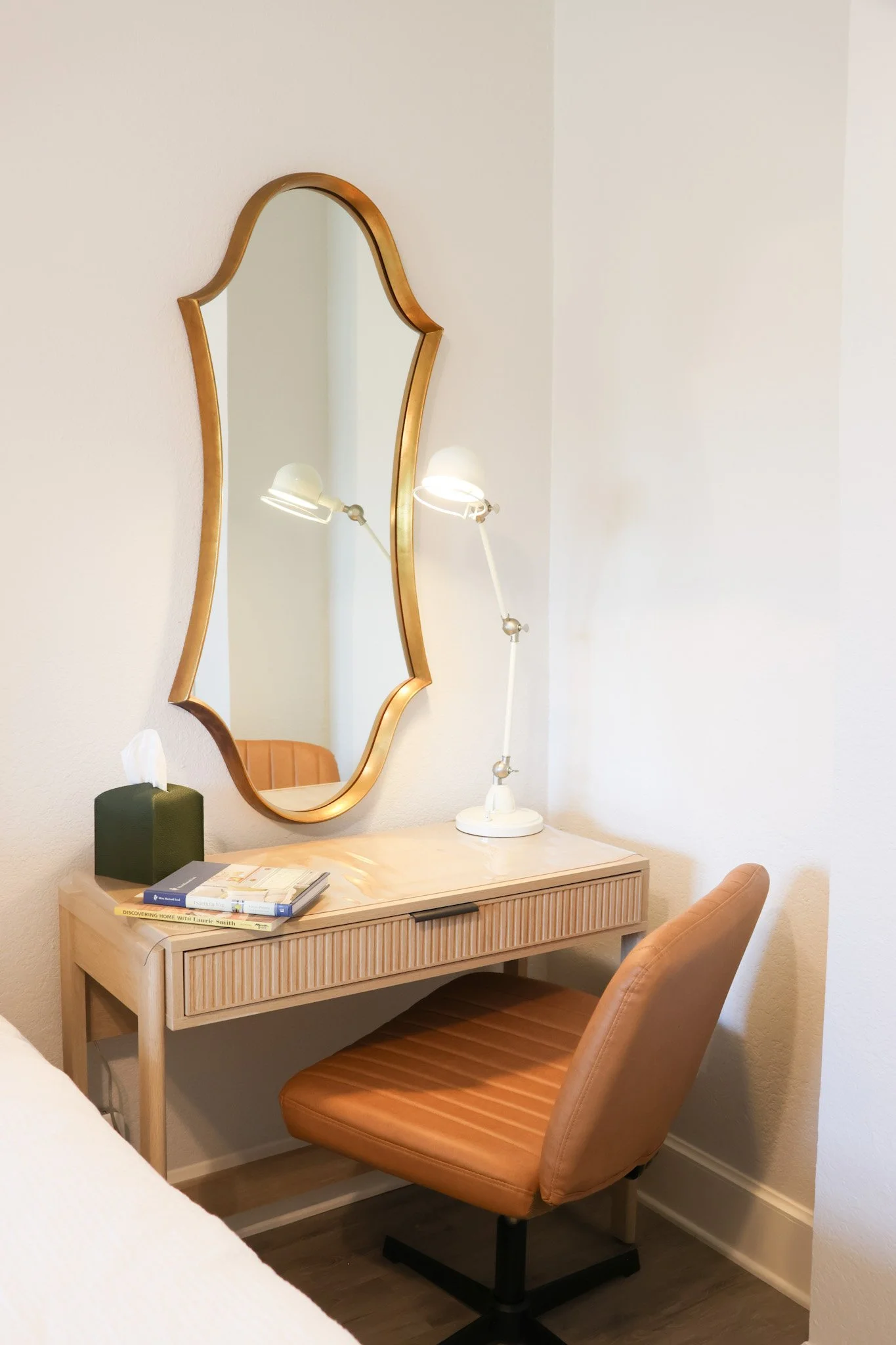 A wooden desk with a tan upholstered chair, a tall green tissue box, and stacked books on top. A large, wavy-edged gold-framed mirror hangs on the wall above the desk. A white adjustable lamp is on the desk, and part of a beige chair is visible to th